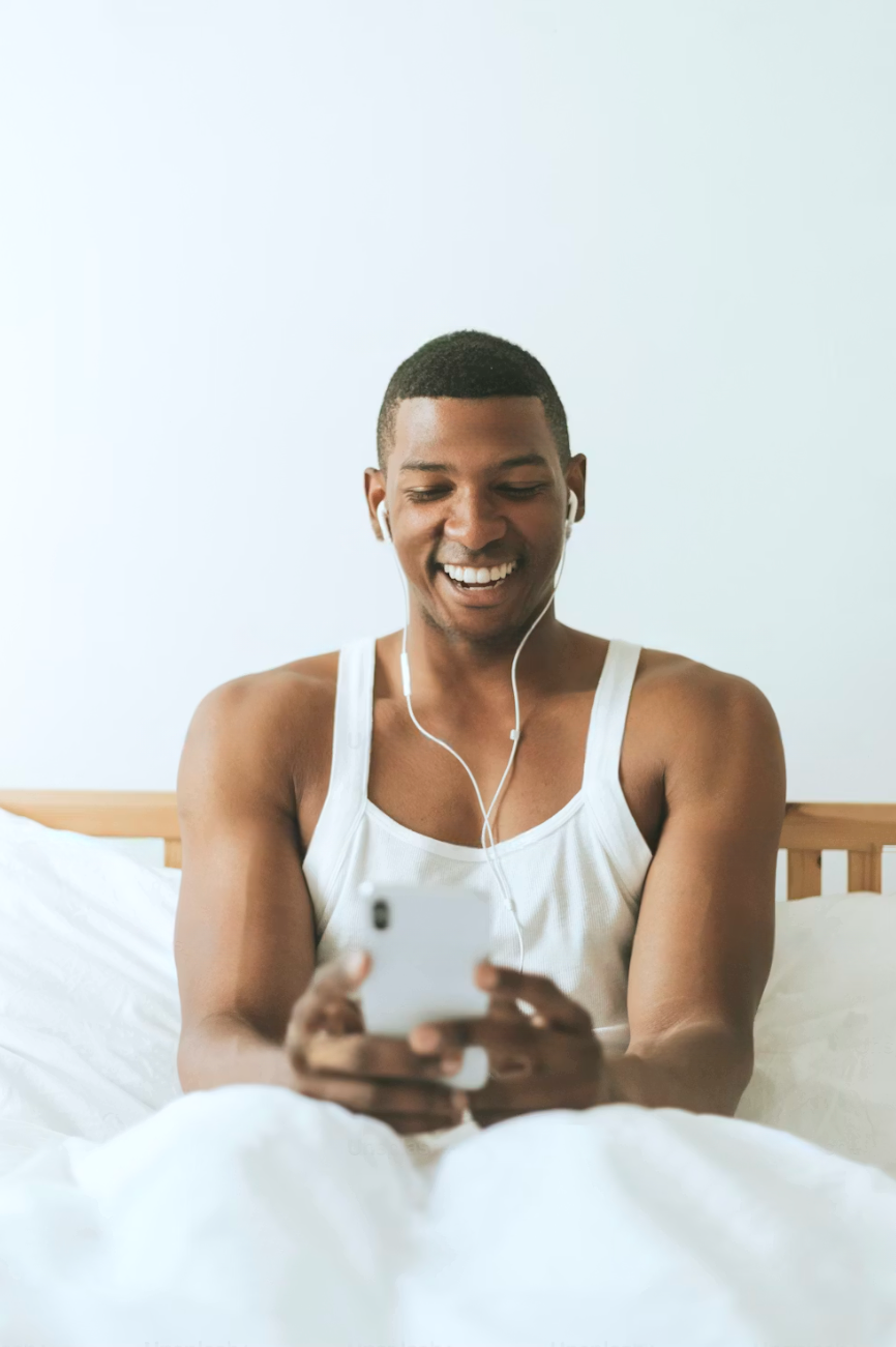 A young man sitting on a bed, smiling while looking at his smartphone, with white earbuds in his ears, wearing a white tank top, and a wooden headboard behind him.