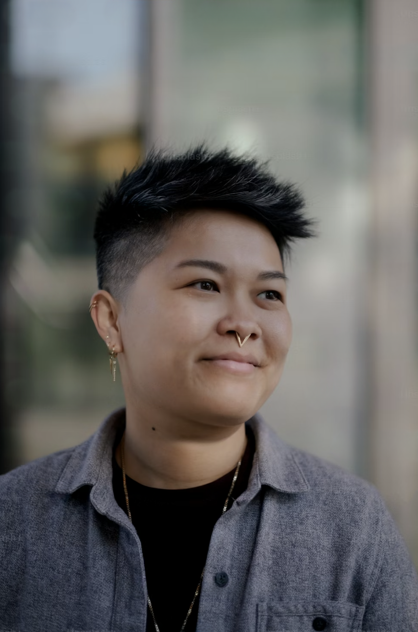 A person with short, spiky dark hair, wearing a septum ring and earrings, smiling slightly while looking to the side, dressed in a gray shirt layered over a black shirt, with a blurred background.