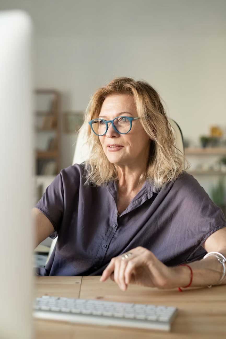 A middle-aged woman with blonde, wavy hair and glasses sitting at a desk, working on a computer in a well-lit room.