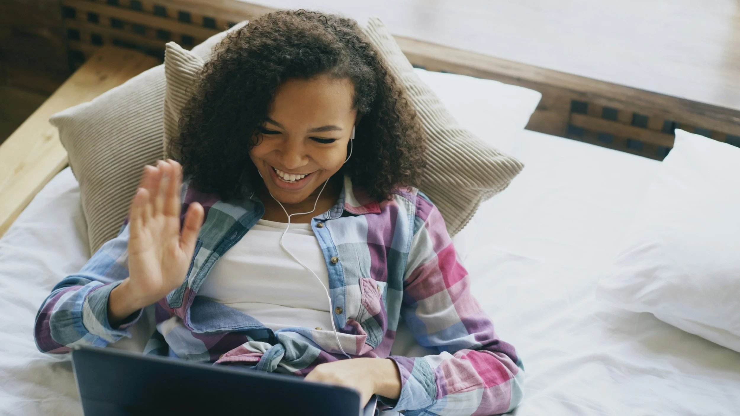 A woman with curly hair is lying on a bed, smiling and waving at a laptop screen while wearing earbuds.