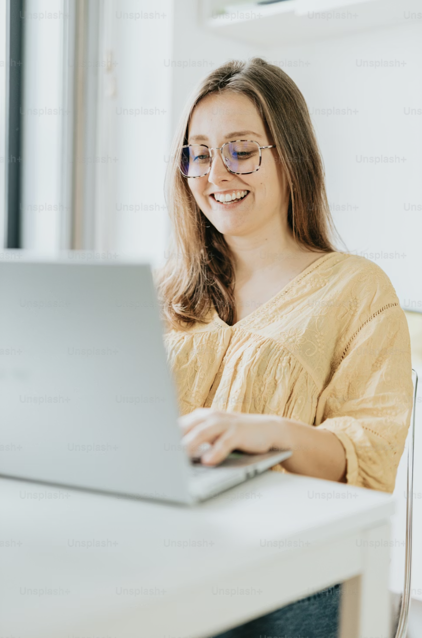 A woman with glasses smiling while working on a laptop at a white desk in a bright room.