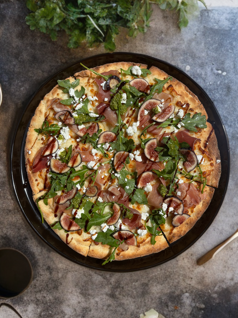 A pizza topped with arugula, figs, prosciutto, blue cheese, balsamic glaze, and sliced almonds.