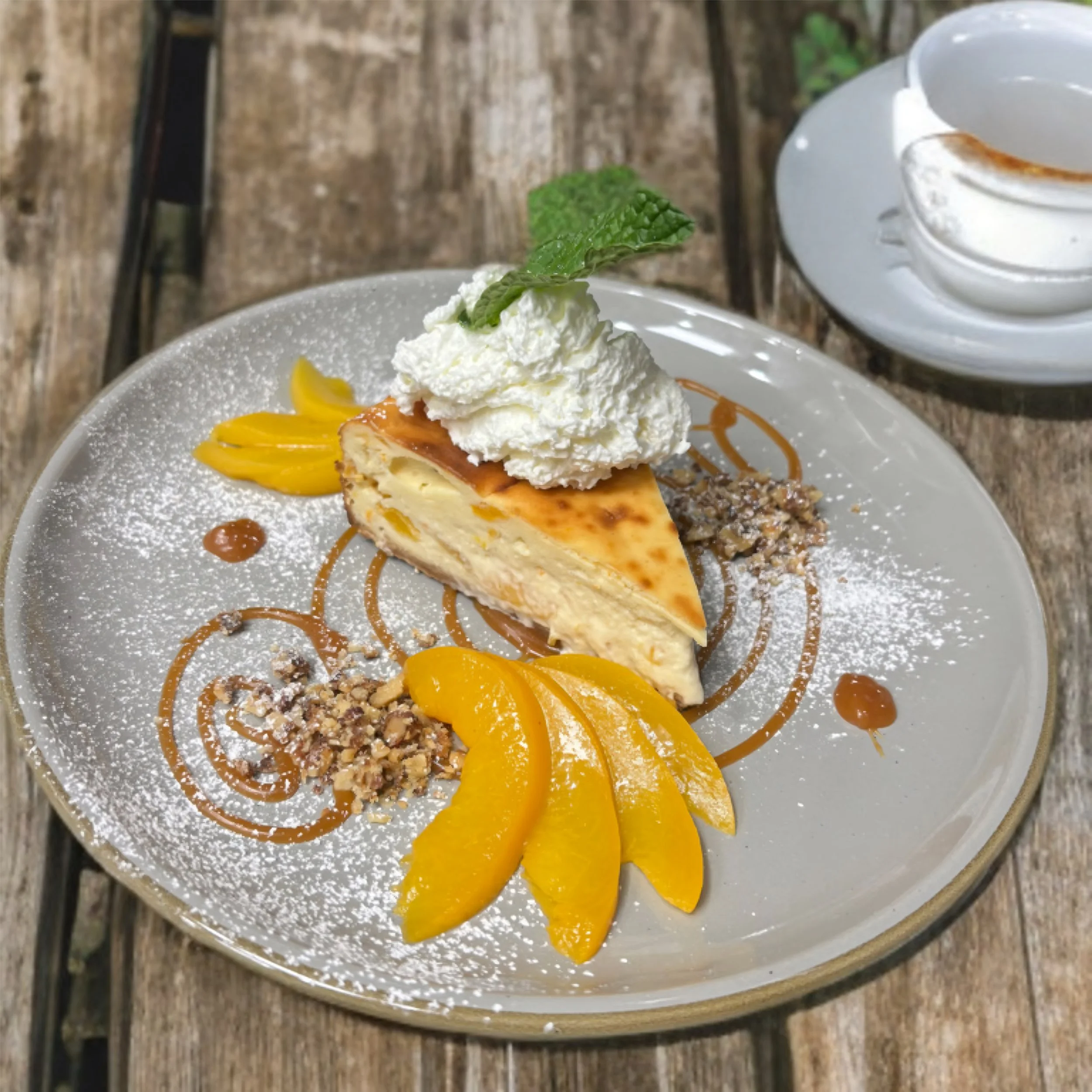 A slice of cheesecake topped with whipped cream and a mint leaf, garnished with peach slices, chopped nuts, caramel drizzle, and a dusting of powdered sugar, on a decorative plate.