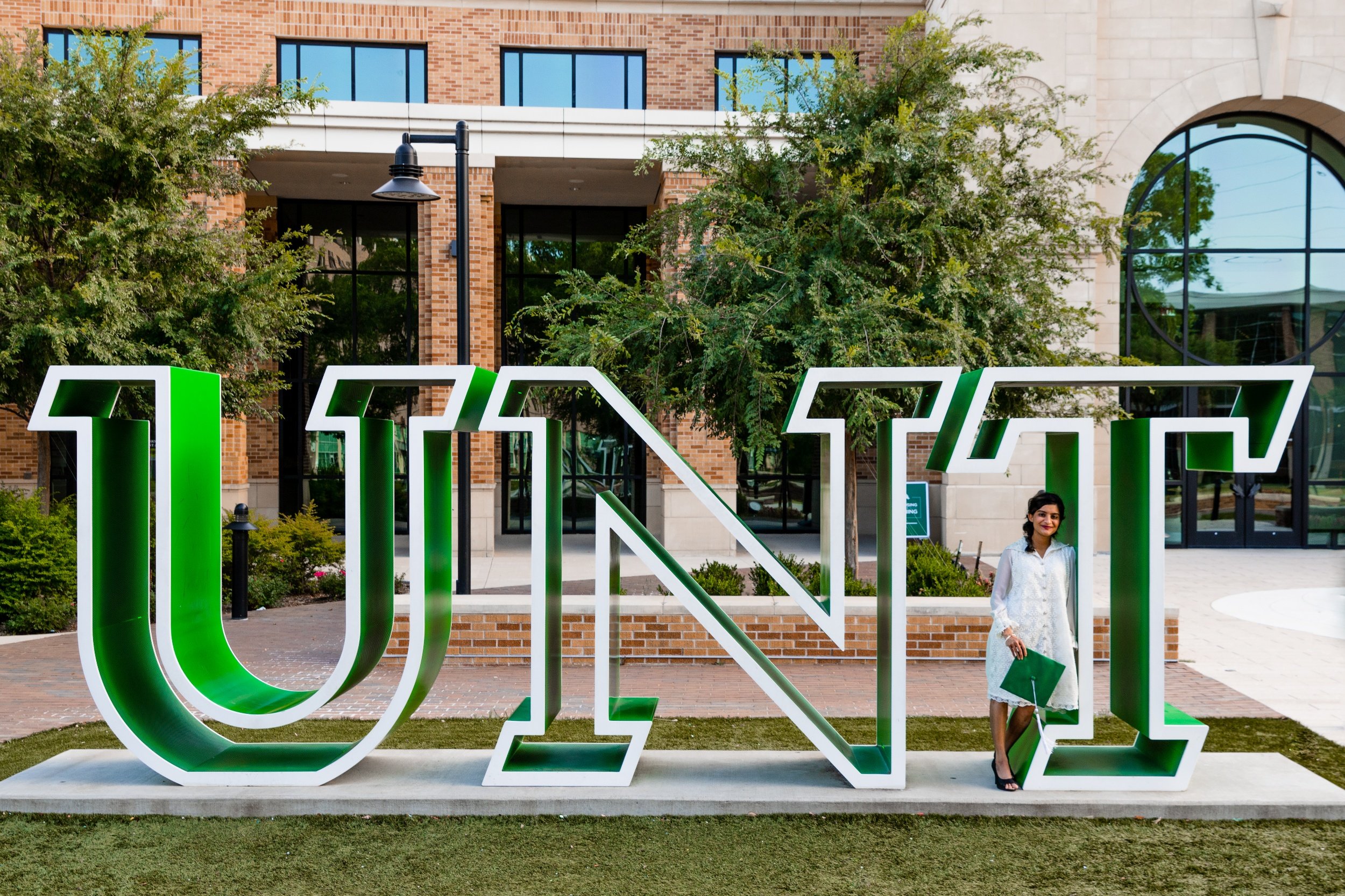 A woman standing inside a large green and white sign spelling 'UINK' outside a modern brick building with glass windows and trees.