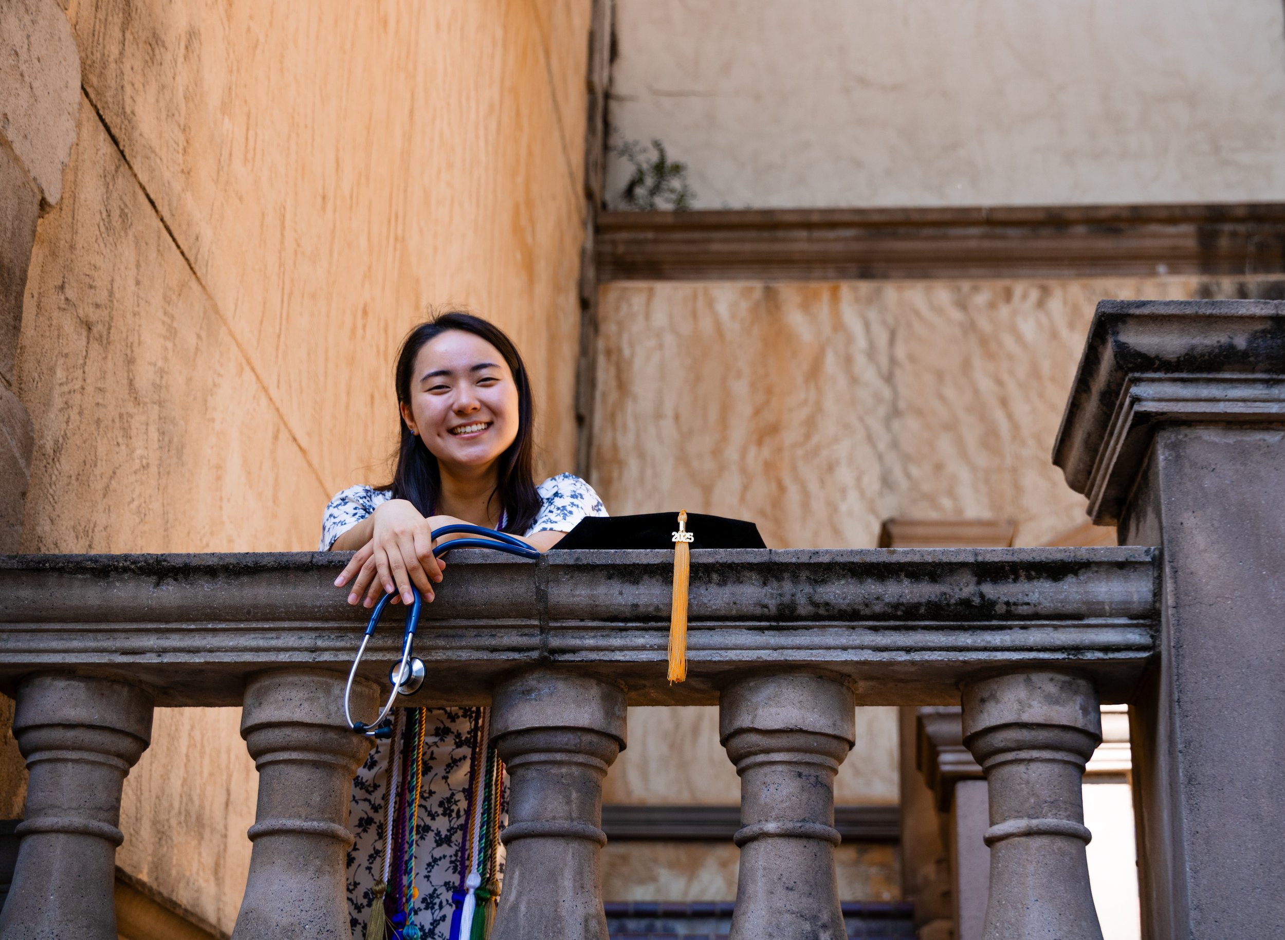 Smiling young woman leaning on a stone balcony railing with her arms resting and graduation cap with tassel on the railing, holding her stethoscope, in an outdoor courtyard setting.