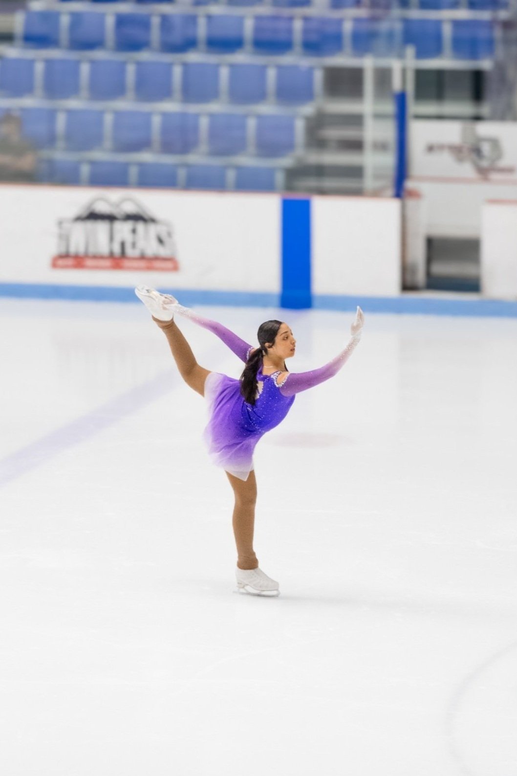 A female figure skater dressed in a purple and white costume performs a move on an ice rink.