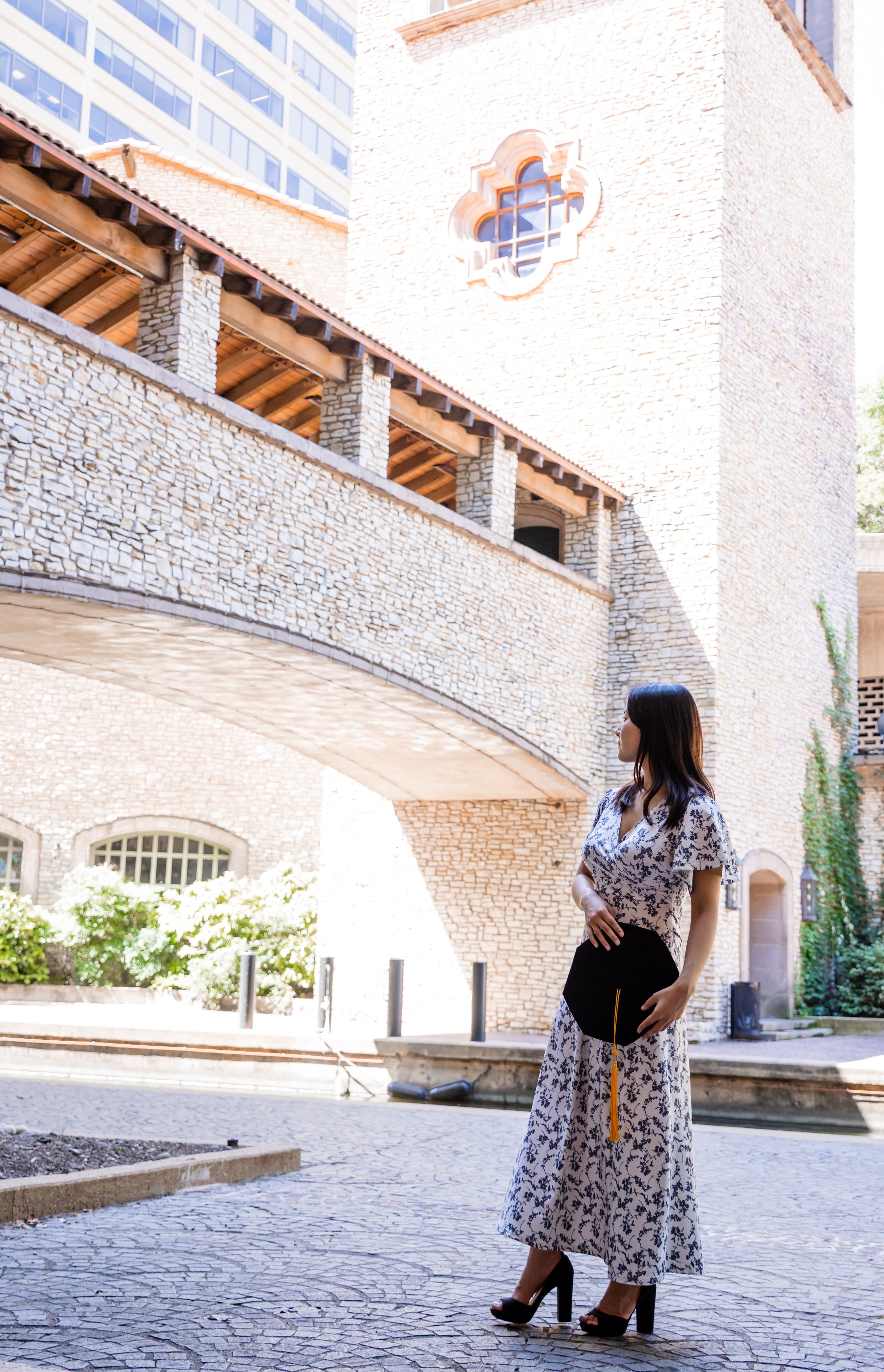 A woman in a floral dress and high heels holding a diploma stands outside near a stone bridge and historic brick building with a decorative window.