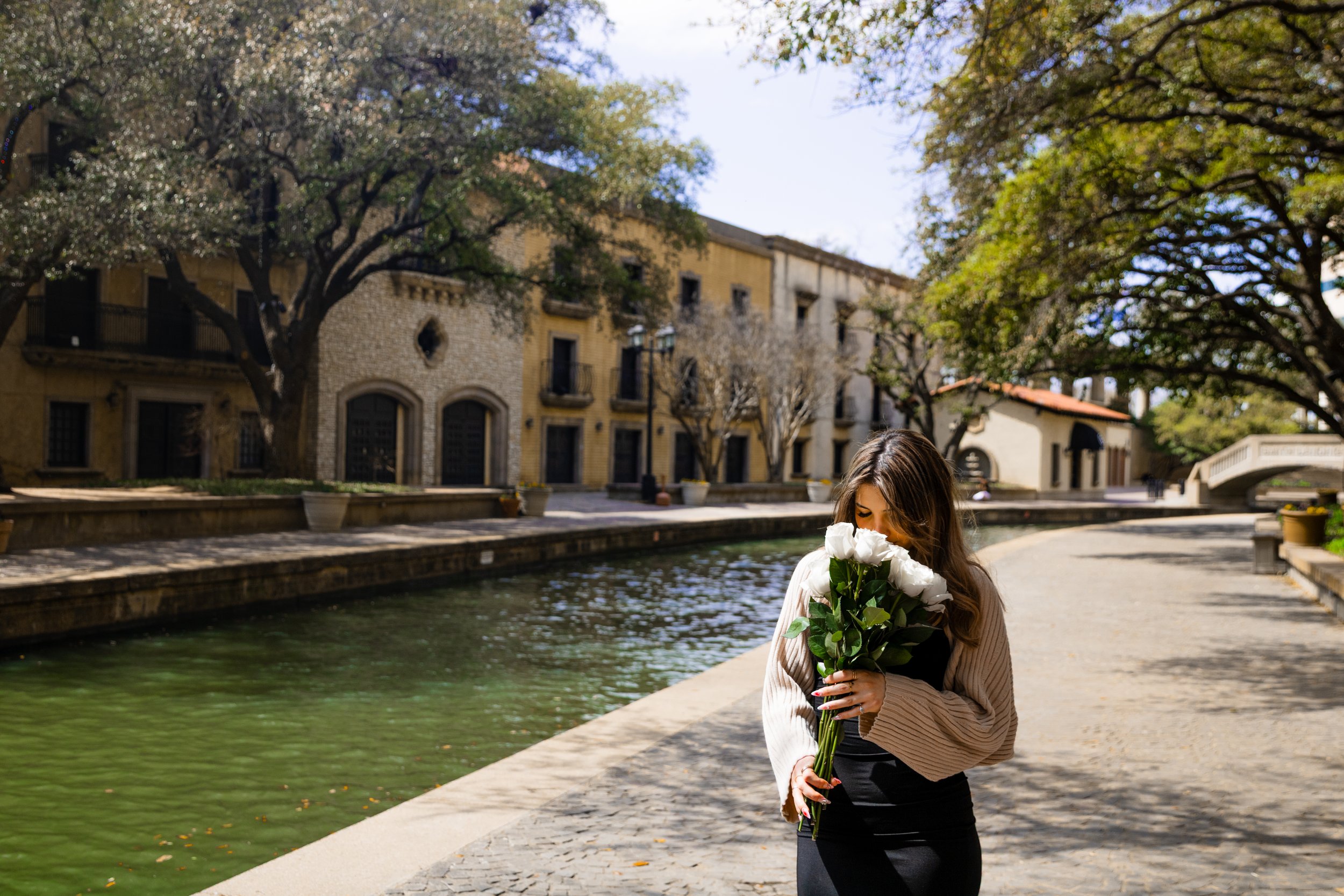 A woman holding a bouquet of white roses on a sunny sidewalk next to a canal with trees and historic buildings in the background.