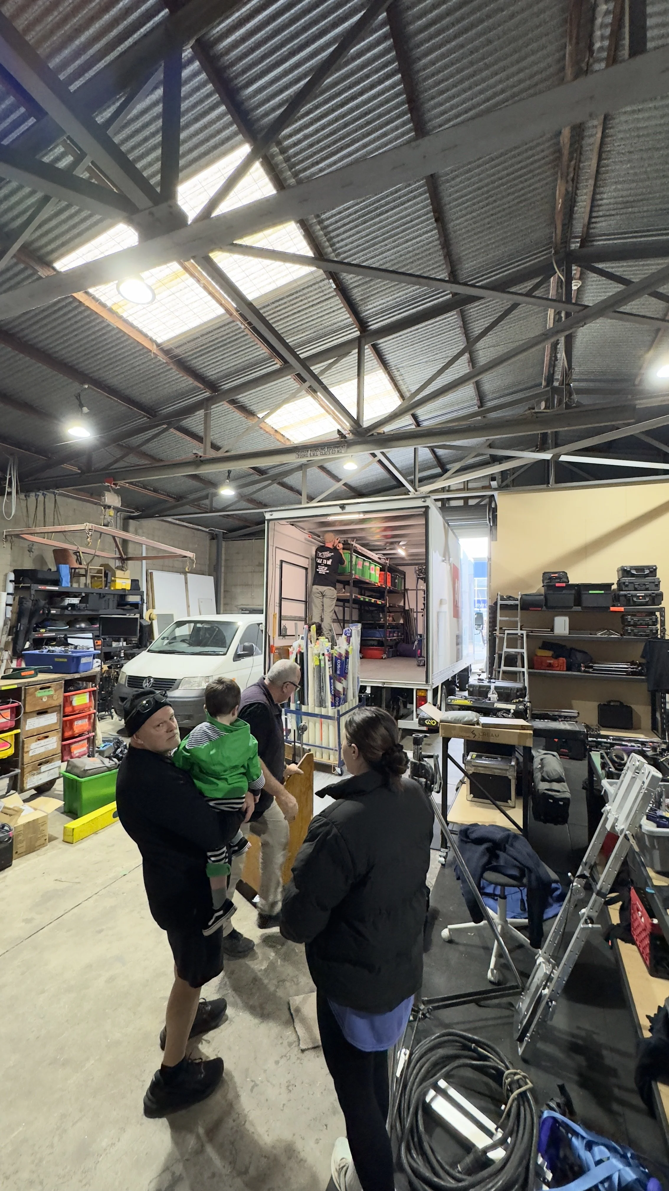 Interior of a workshop or garage with various tools and equipment, a parked van, and people interacting near a partially open truck containing more equipment.