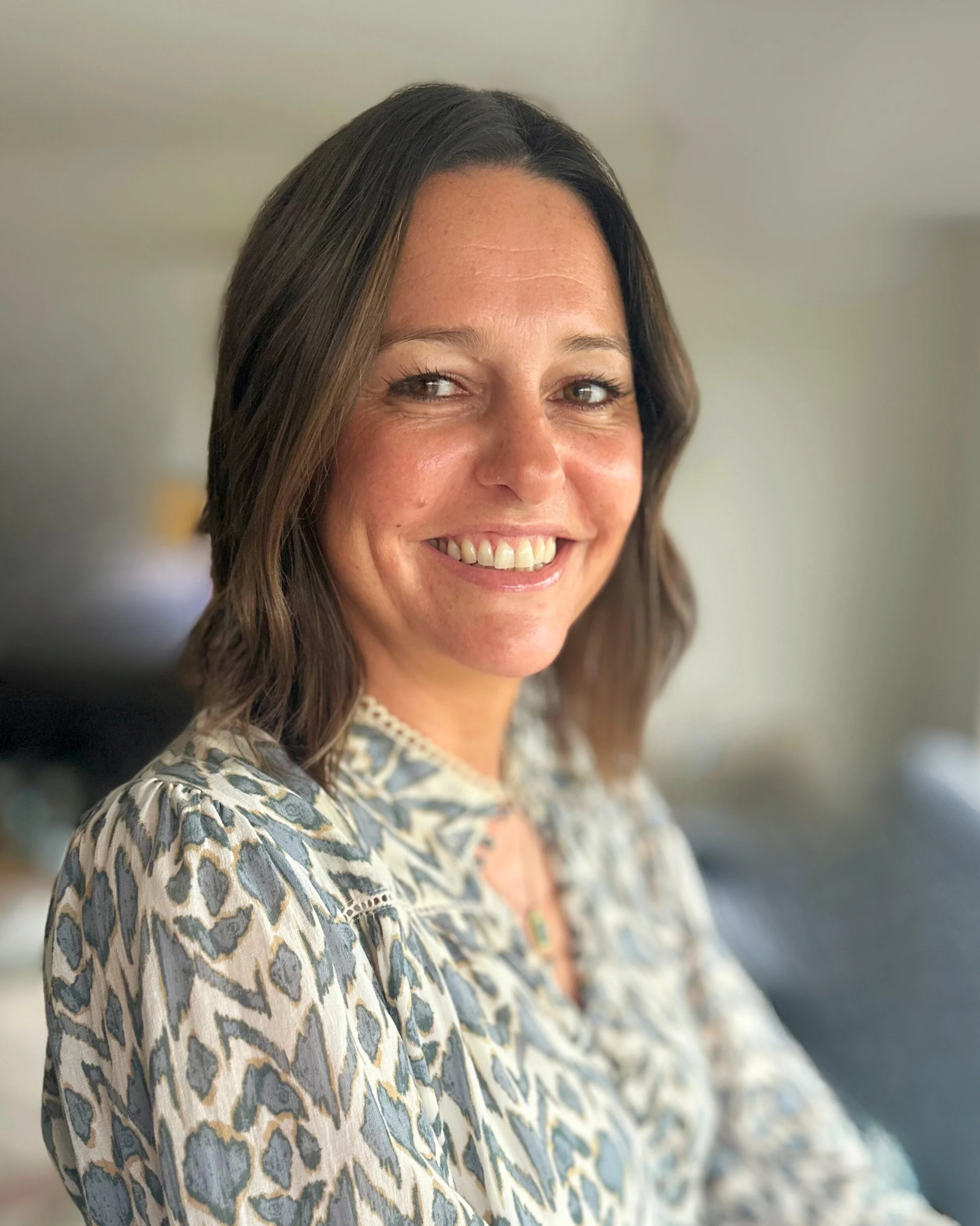 A woman with shoulder-length brown hair smiling, wearing a patterned blouse, in a softly lit room.