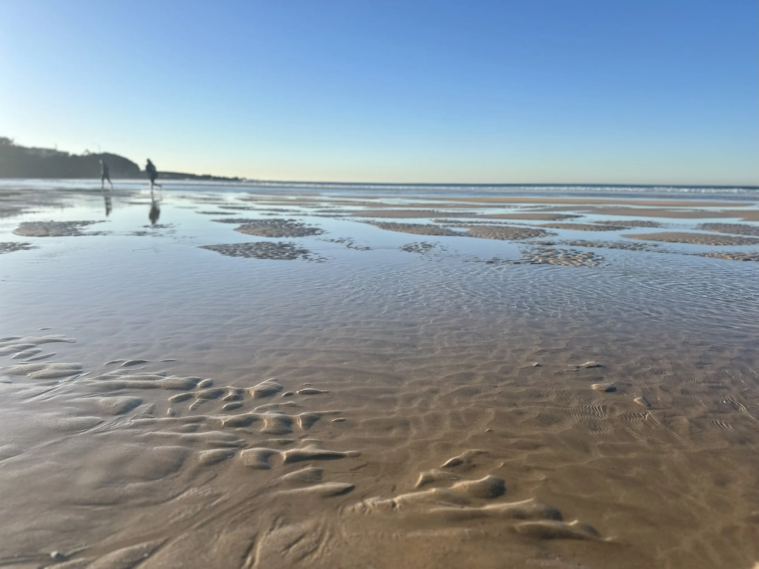 Empty sandy beach with shallow water and footprints, two people walking in the distance, blue sky, calm ocean, and distant landmass.