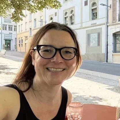 A smiling woman looking at the camera, sitting in a cafe underneath a tree on a summer day