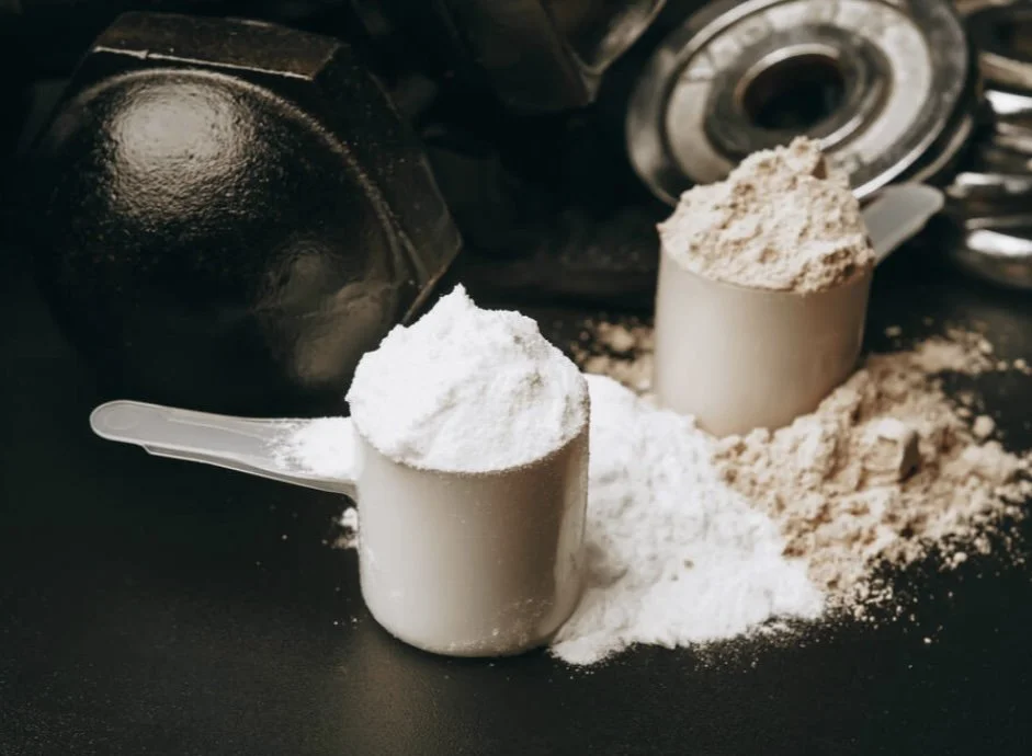 Scoops of protein powder or supplement powder in white measuring cups surrounded by additional powder on a dark surface with fitness equipment in the background.