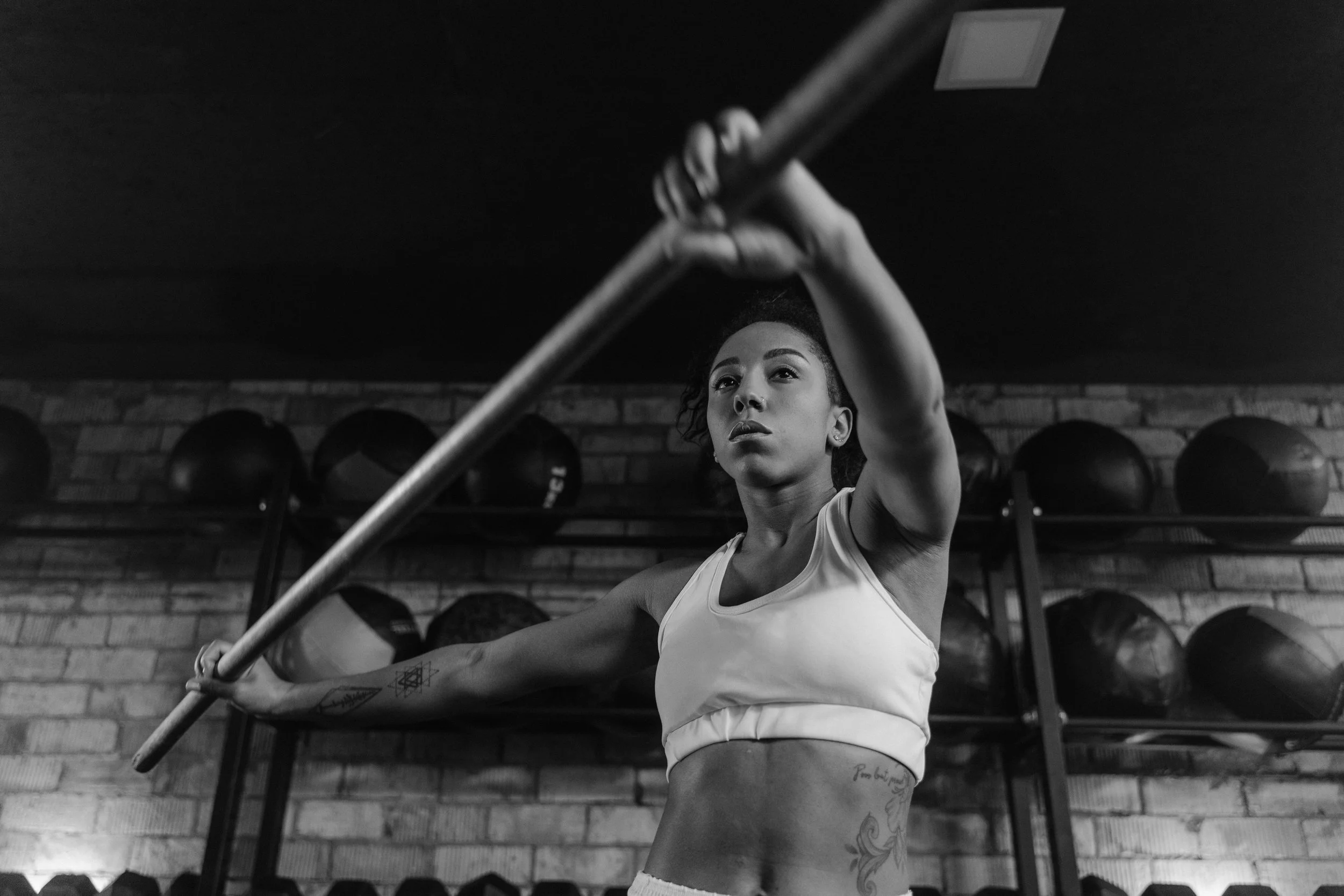 A woman lifting a barbell in a gym with brick walls and weight balls in the background.