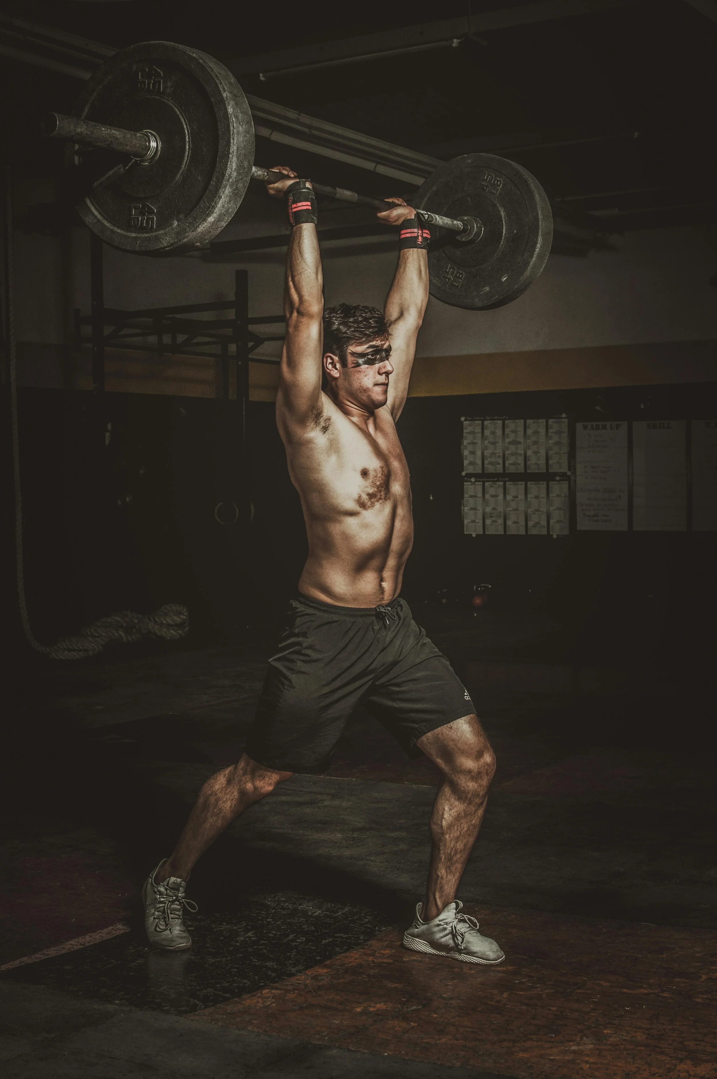 A shirtless man with face paint, wearing black shorts and sneakers, lifting a barbell overhead during a workout in a gym.
