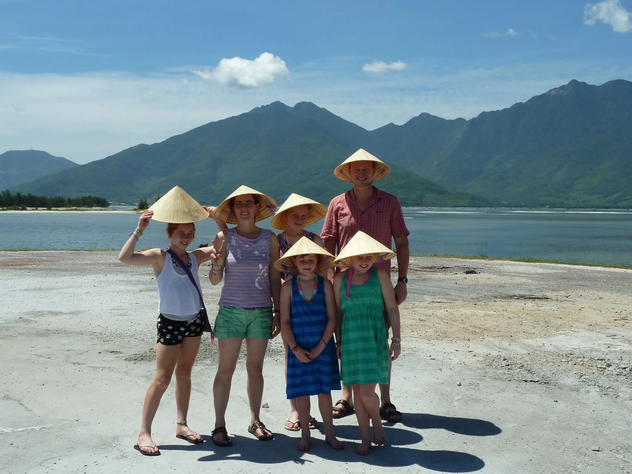 Family wearing conical hats posing by a scenic lake with mountains in the background on a sunny day.