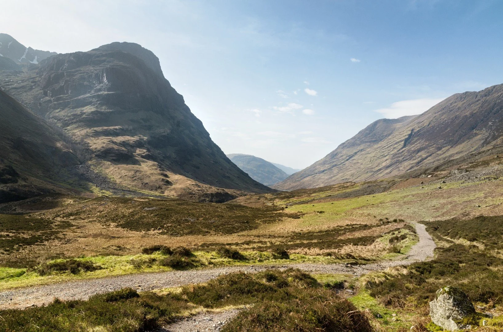 Scenic view of a mountainous landscape with a winding path, green hills, and rocky peaks under a clear blue sky.