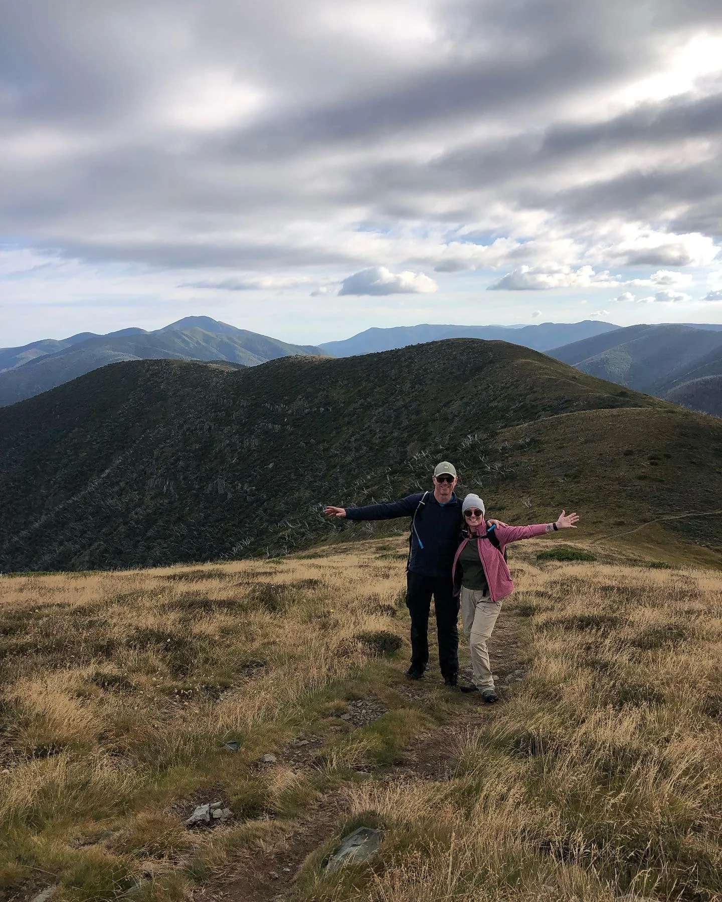 Two people hiking on a mountain trail with expansive views of distant hills and a cloudy sky.