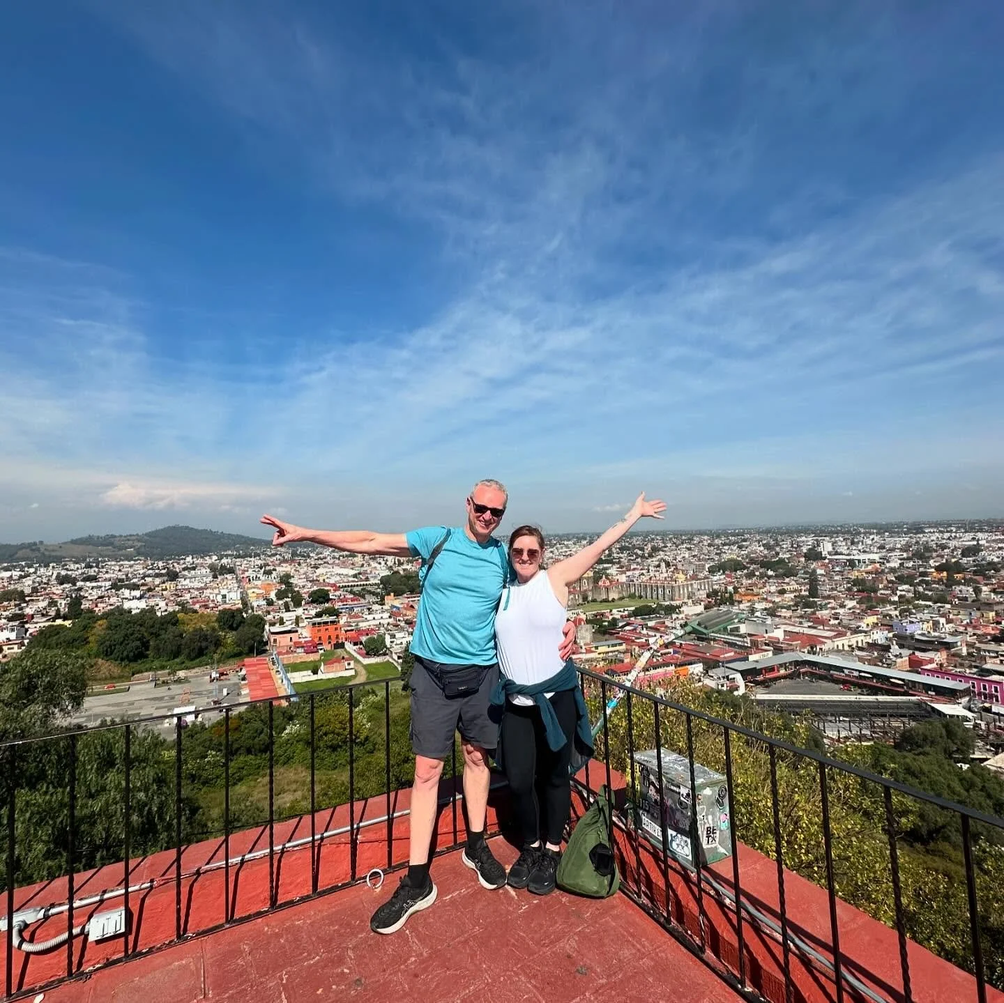 A man and woman standing on a rooftop with arms outstretched, overlooking a scenic cityscape on a clear day.