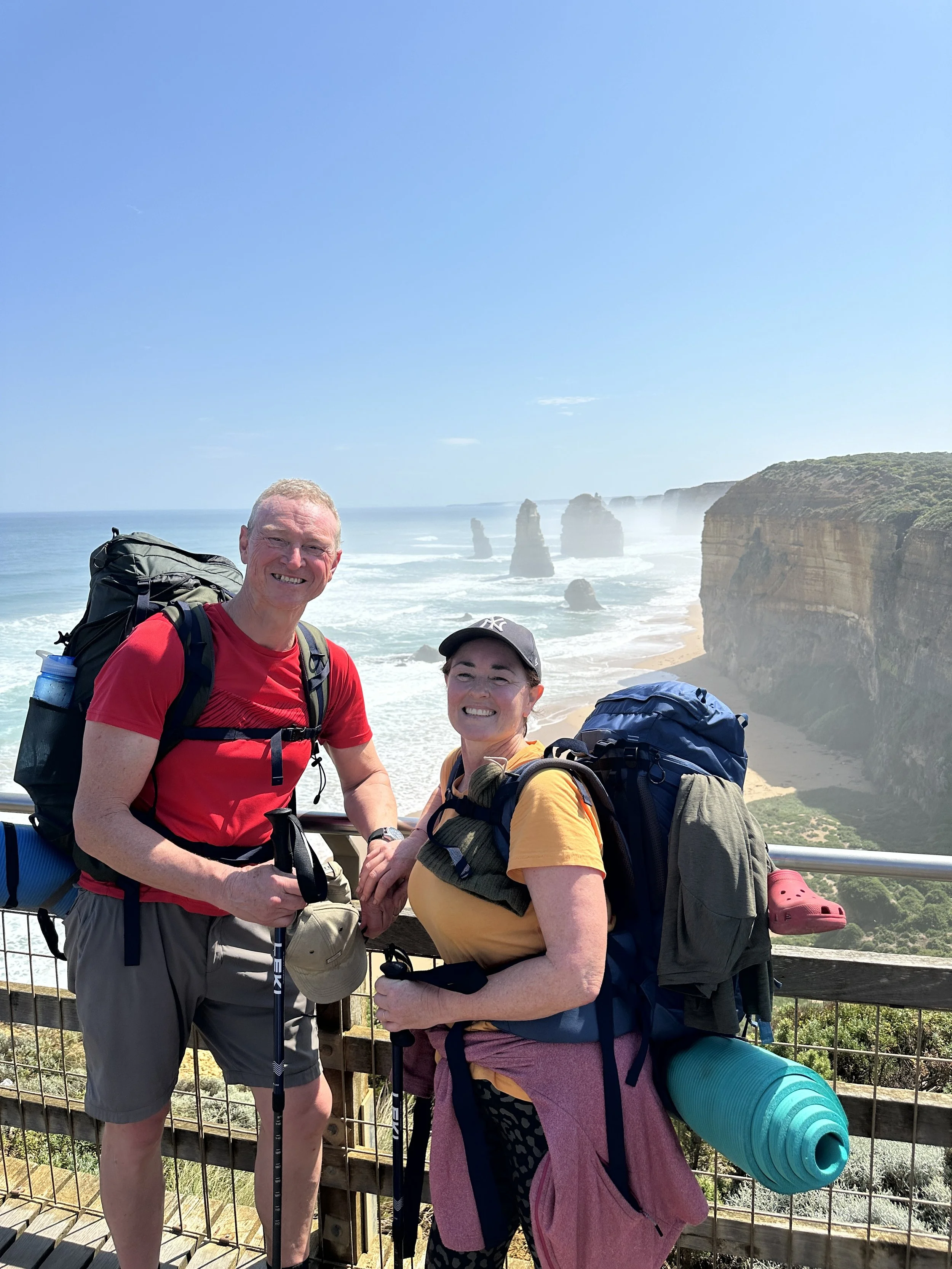 Two hikers with backpacks standing on a viewing platform overlooking the ocean and rock formations, known as the Twelve Apostles, on a sunny day.