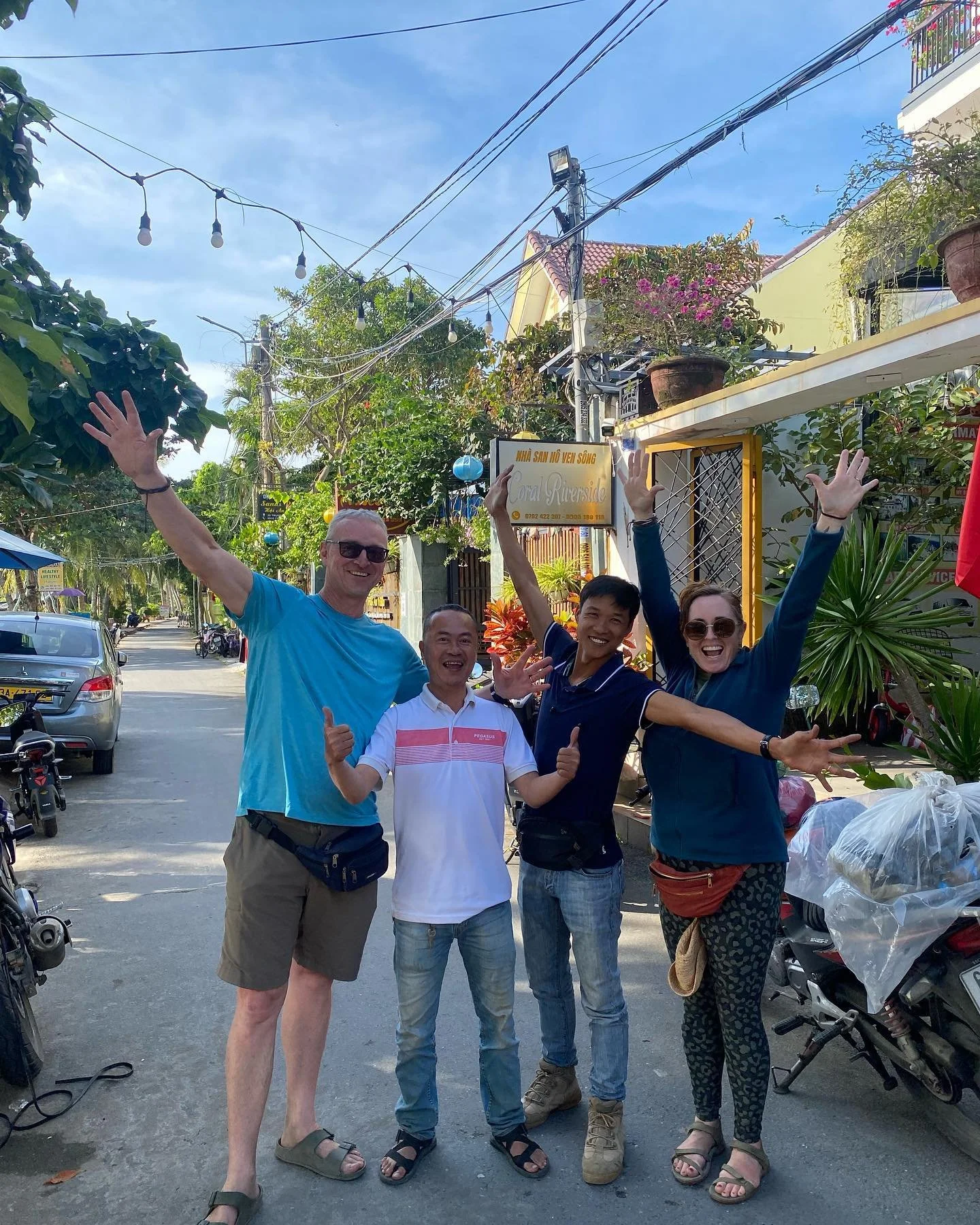 Four people smiling and posing with arms raised in a street setting, next to a sign that says "Rural Riverside." The background shows potted plants, motorbikes, and buildings.
