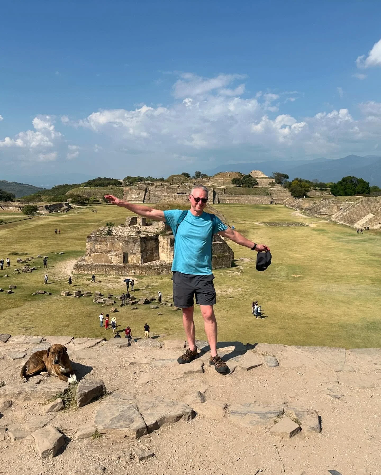 A man in a blue shirt standing with arms outstretched on ancient ruins at Monte Albán, Oaxaca, with a dog sitting nearby and people exploring the site in the background.