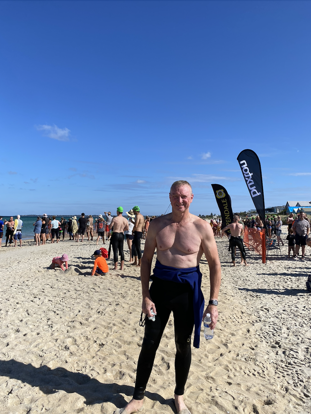 Man in wetsuit standing on a crowded sandy beach with people in swimwear and flags in the background under a clear blue sky.