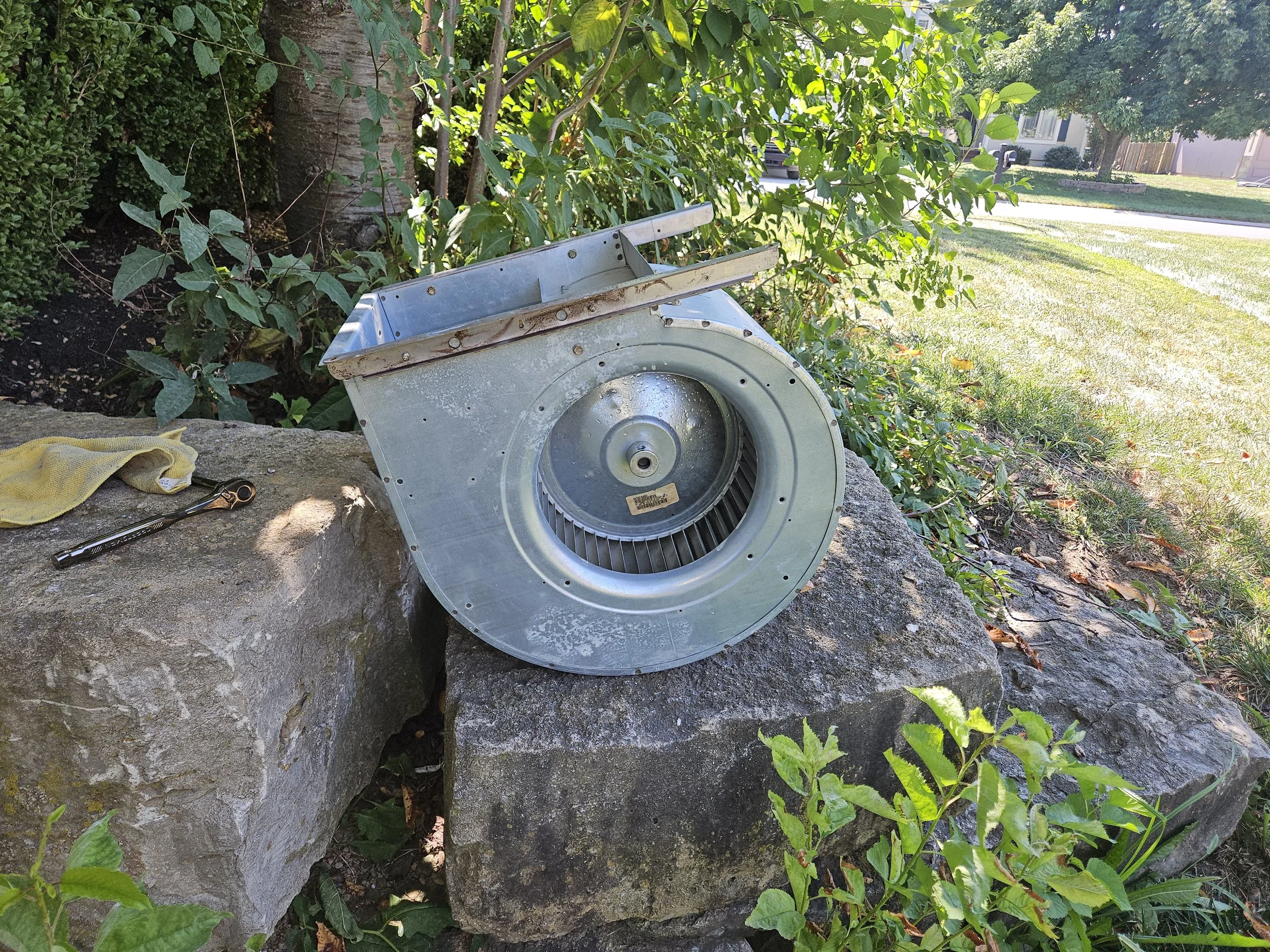 An outdoor scene with a metallic HVAC fan blower assembly placed on large rocks, surrounded by green plants, with a house and street visible in the background.
