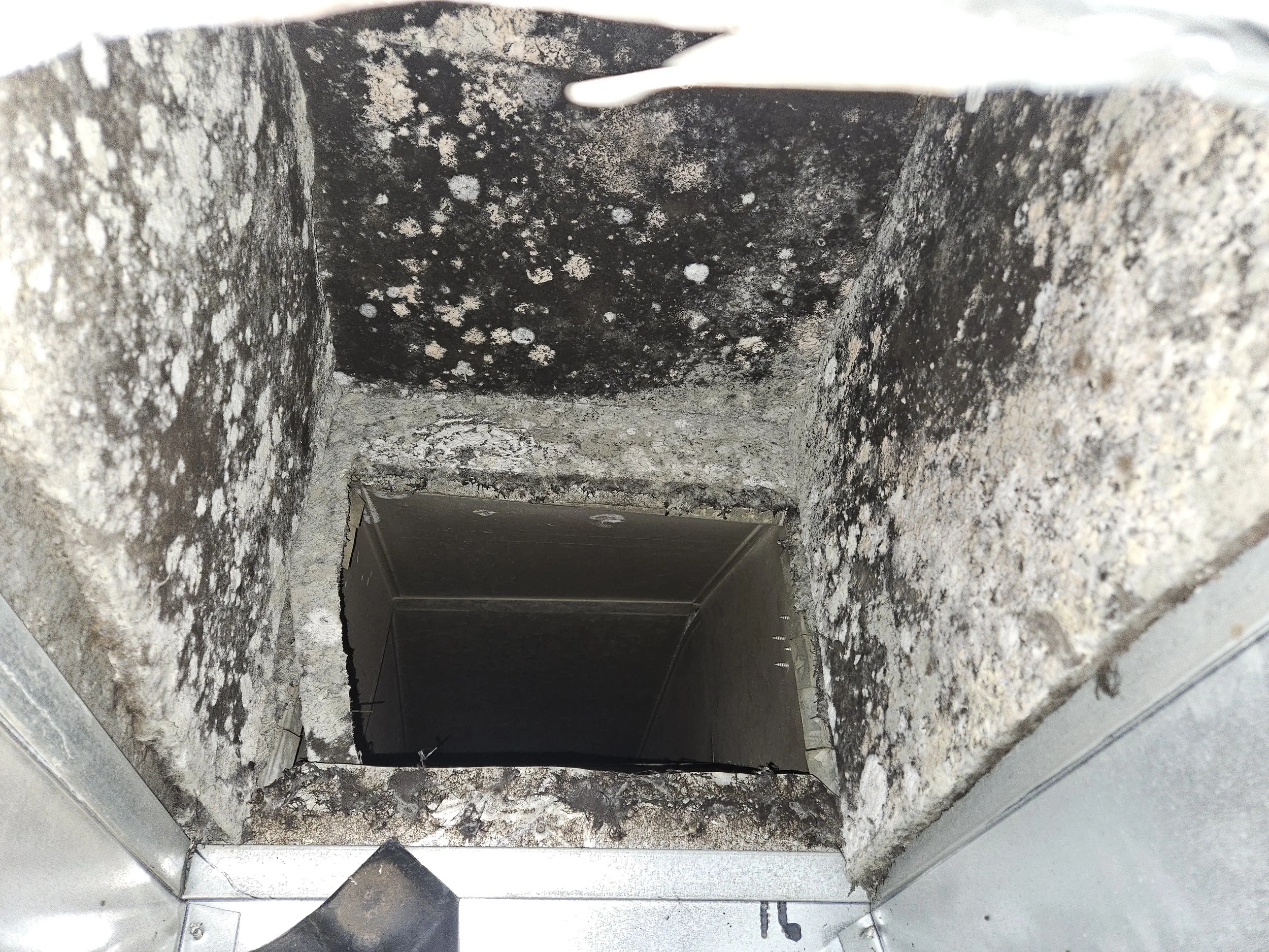 View looking down into a square concrete or metal duct or vent, showing black soot and white mold or mineral deposits inside.