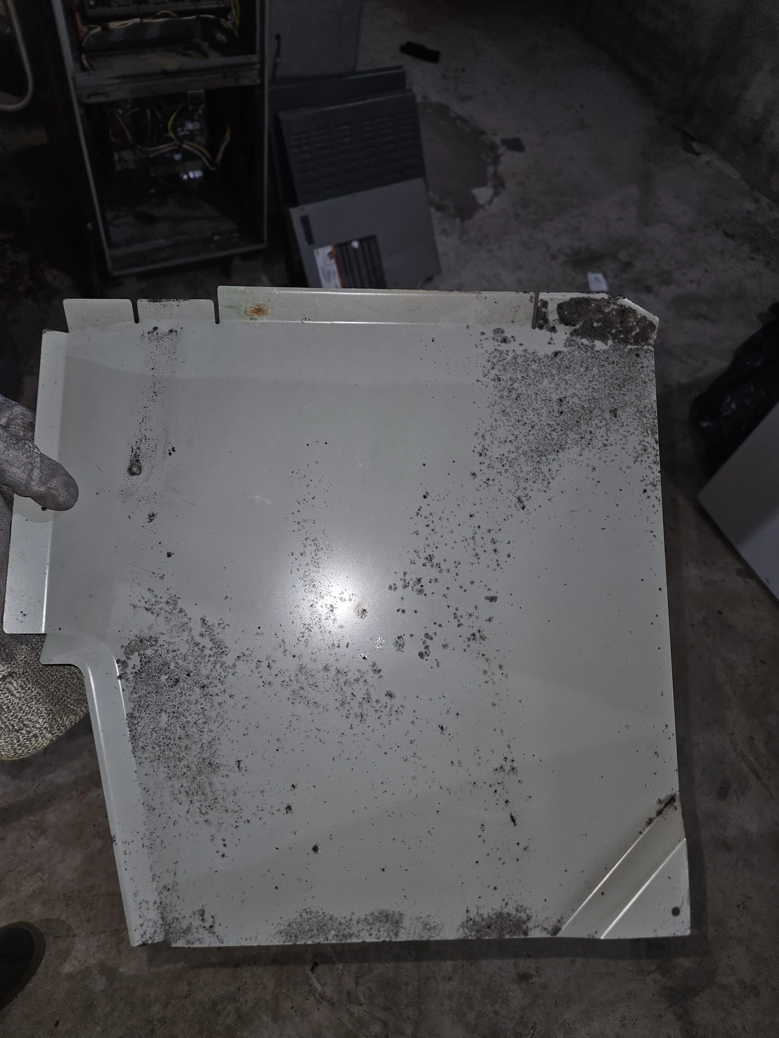A white metal panel with black dust and dirt, held by a person wearing a glove, with other equipment and a concrete surface in the background.