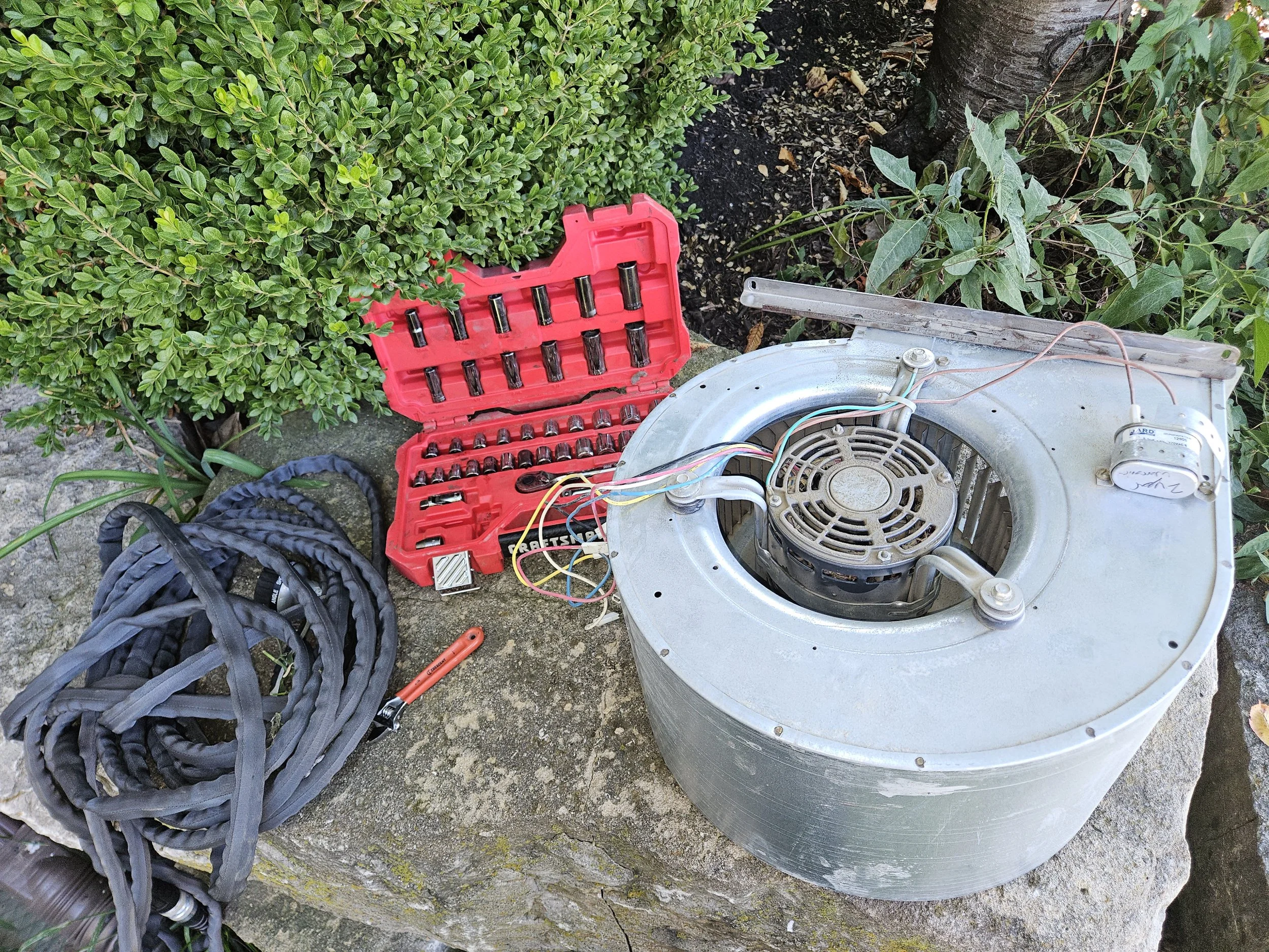 An outdoor scene with a laundry or industrial fan, a red toolkit with various screwdriver bits, a coiled fabric hose or belt, and some wiring, all placed on a stone surface next to green bushes and plants.