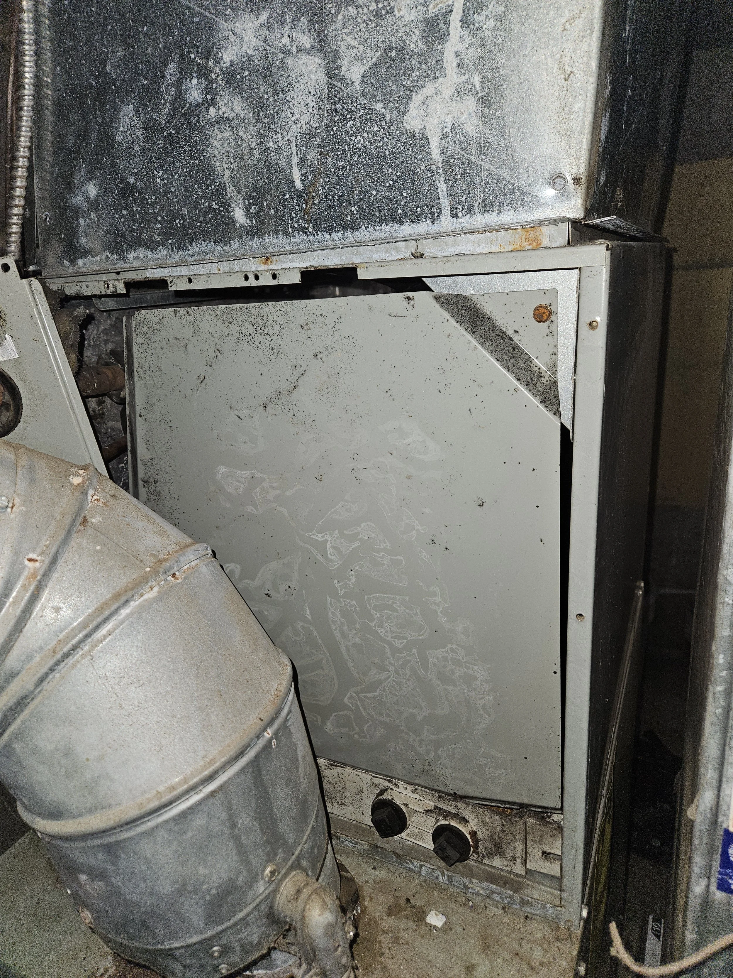 Old, rusty, and dirty furnace with blackened and stained surfaces, with two control knobs at the bottom and an attached large metal pipe on the side.