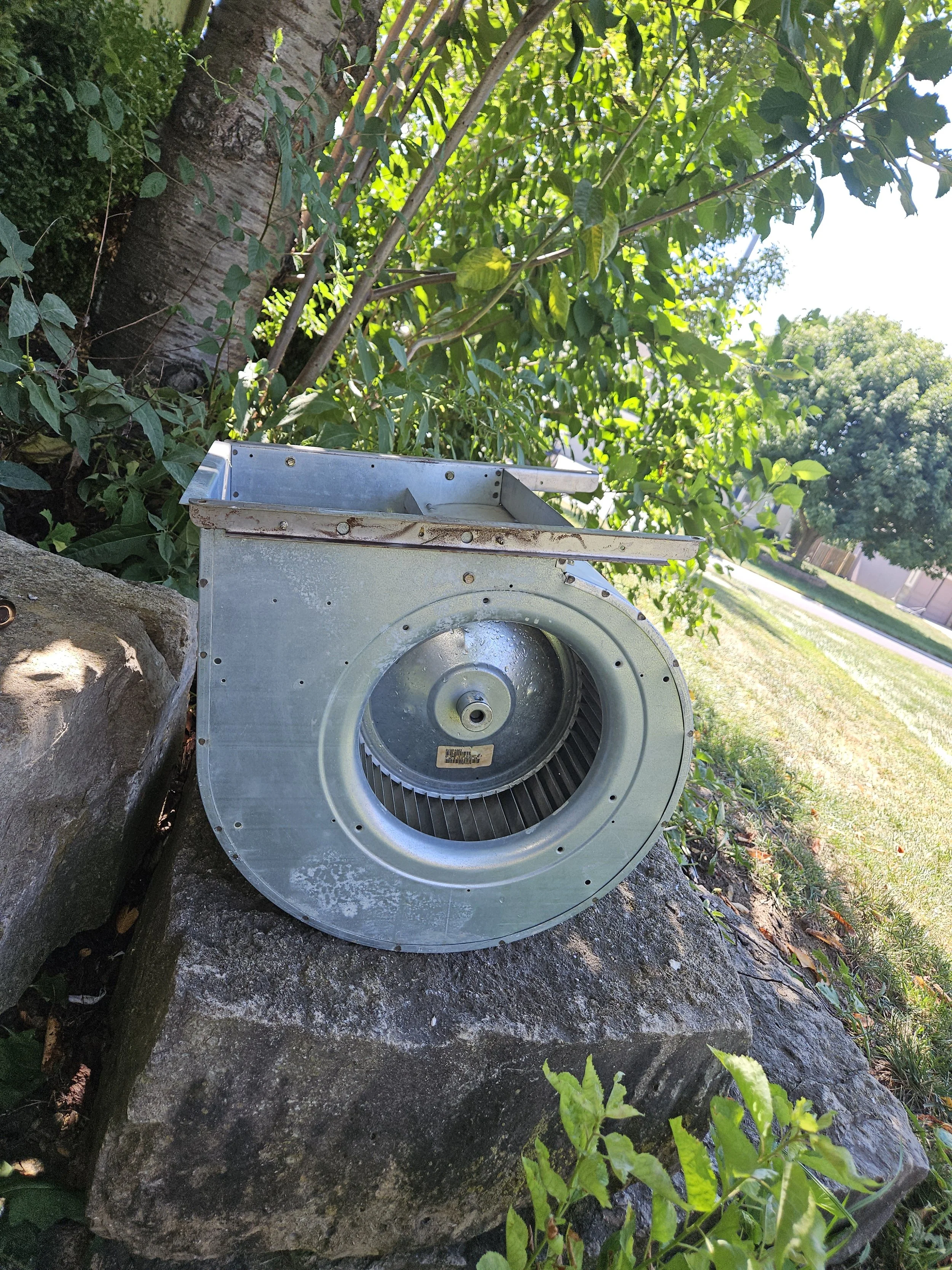 An outdoor scene featuring a metallic HVAC fan blower assembly resting on a large rock next to a tree and green shrubbery.