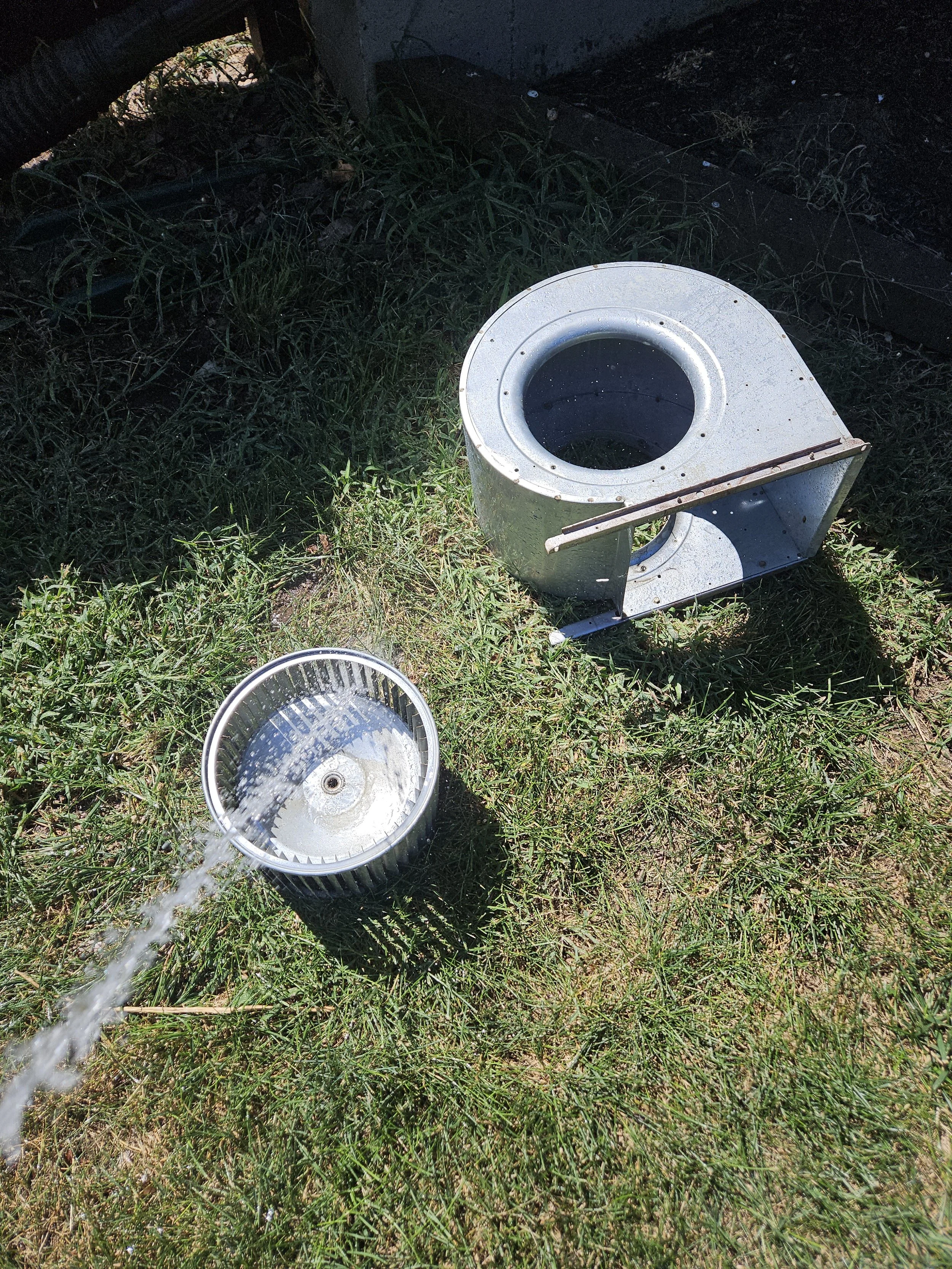 Metal scrap parts, including a round duct and a vent, are on grass with a stream of water pouring into the vent.