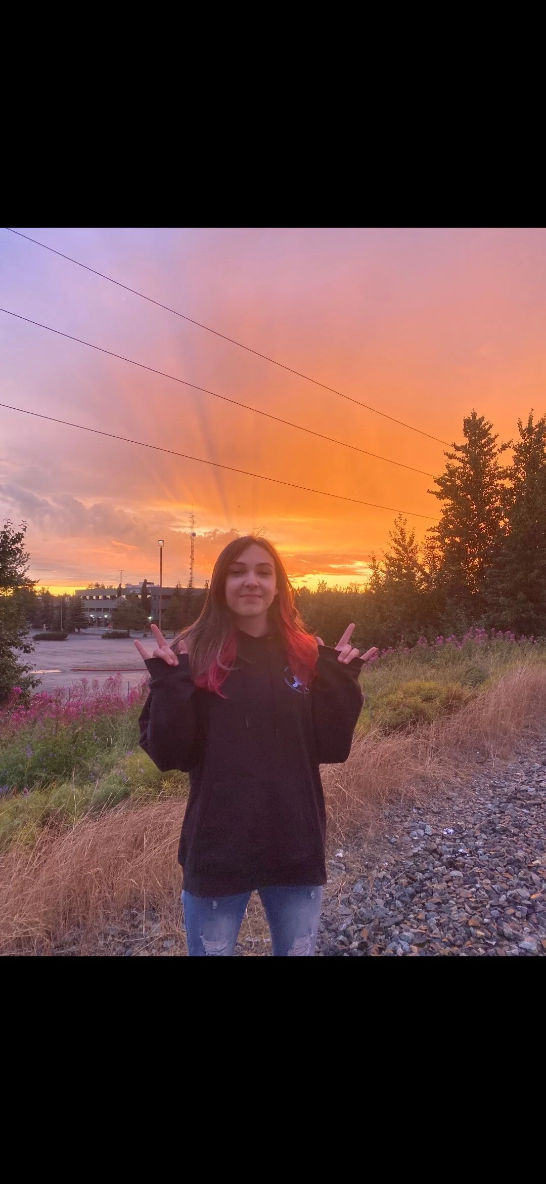 A young woman with brown hair dyed pink at the ends, wearing a black hoodie and ripped jeans, standing outdoors during sunset, making the rock and roll hand gesture with both hands.