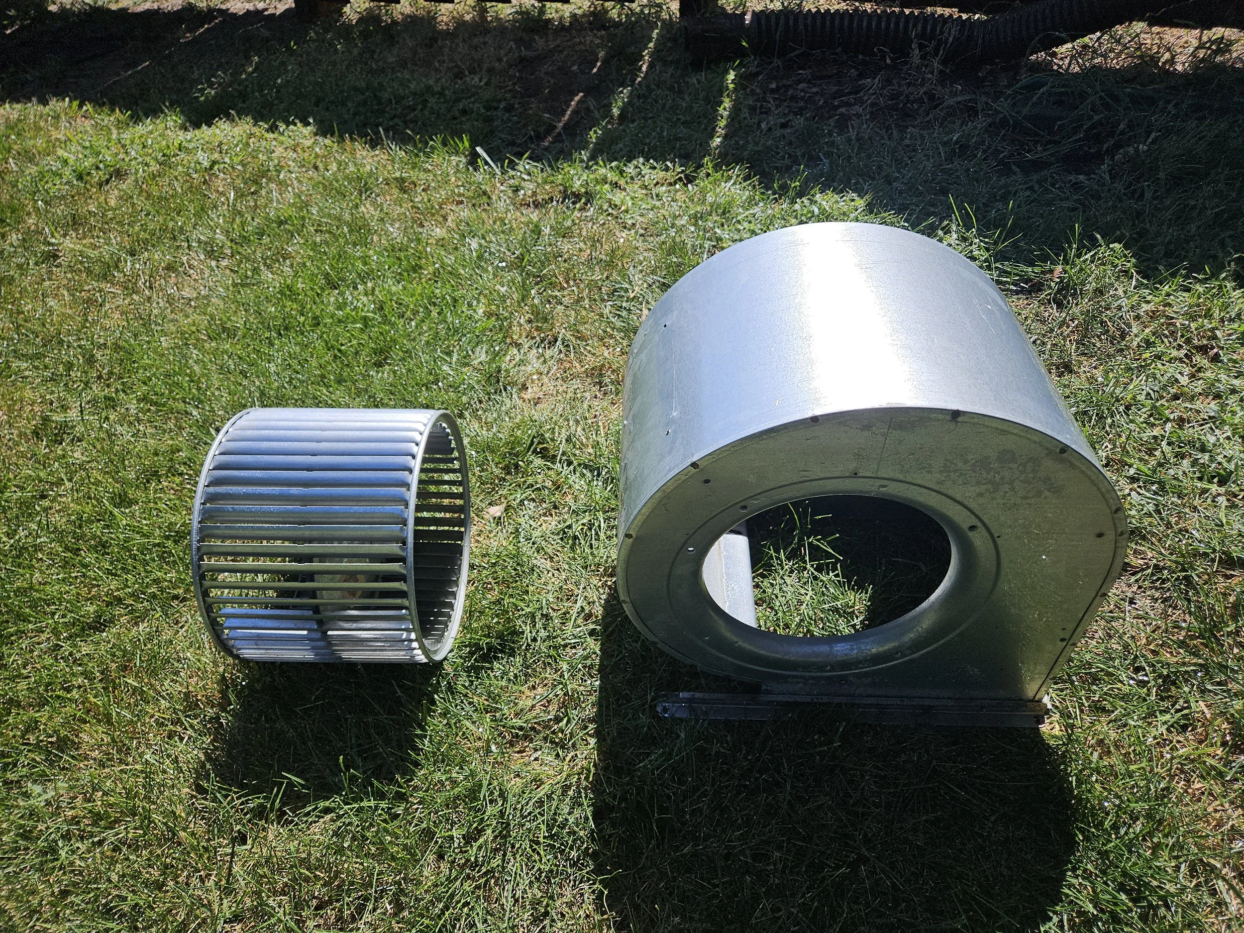 Metal parts, a cylindrical vent or blower, and a fan cage lying on grass outdoors.