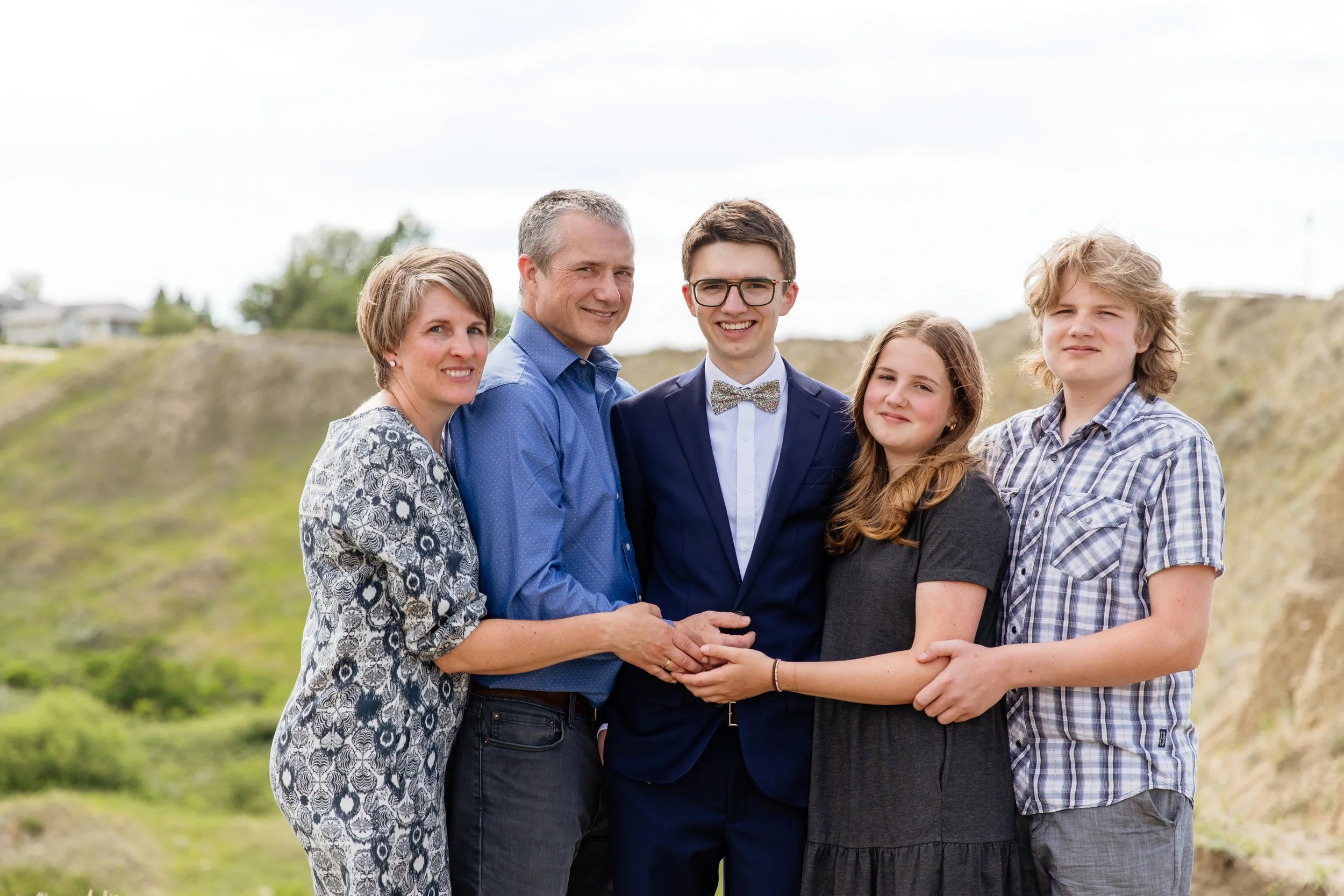 Family of five standing outdoors, holding hands, with a grassy hillside background. The family includes a mother, father, teenage son in a suit, teenage daughter, and young son. They are dressed in casual and semi-formal clothing.