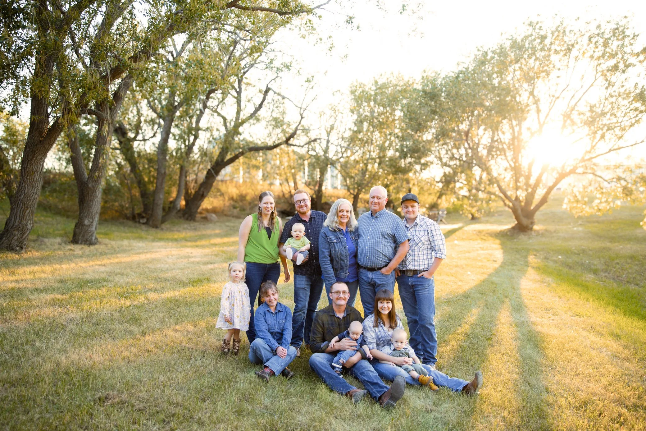 A large family group outdoors in a grassy park during sunset, with trees in the background. The family includes adults and children, some sitting and some standing, all smiling and dressed casually.