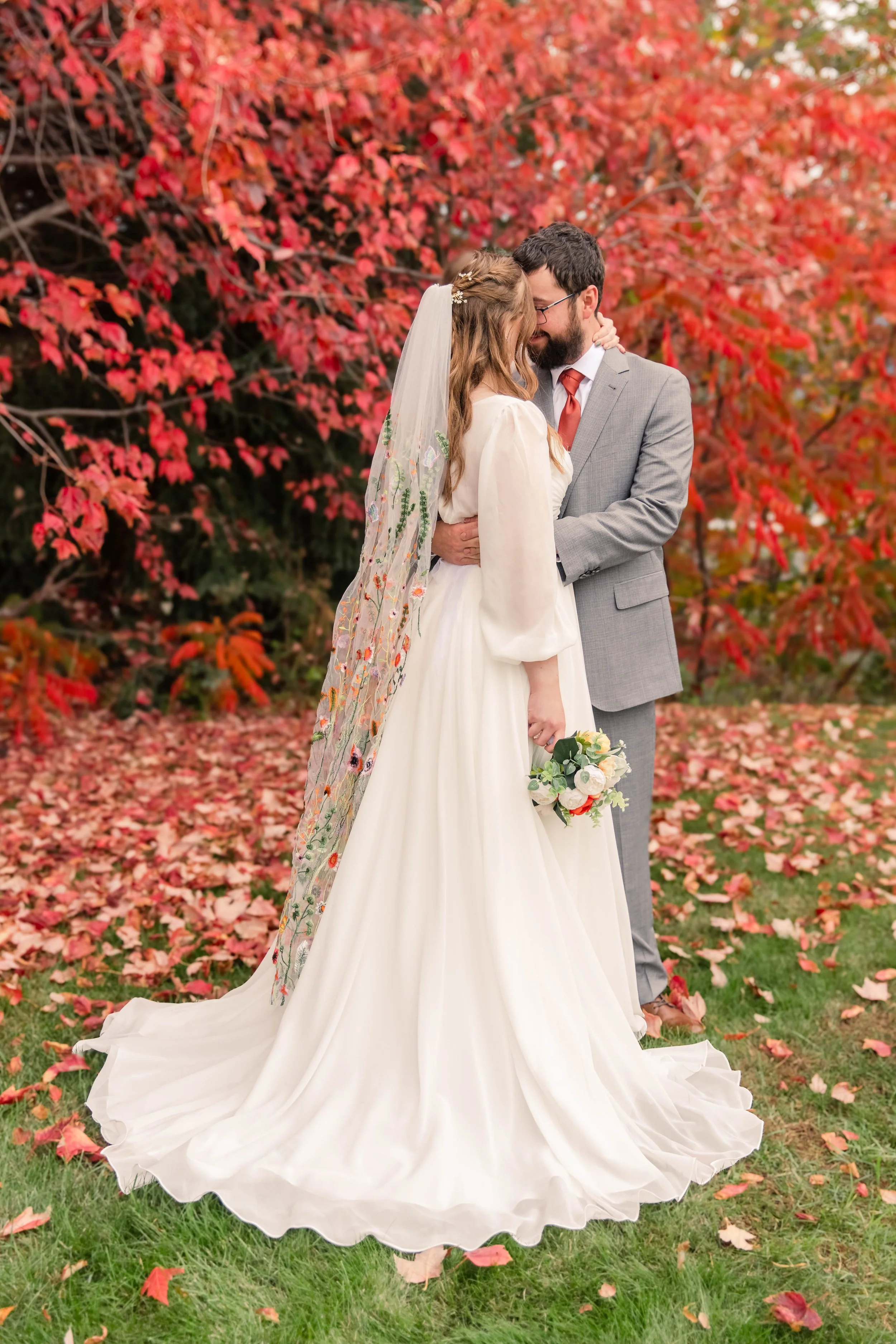 A bride and groom share a kiss on their wedding day outdoors, surrounded by vibrant red and pink autumn leaves, with the bride holding a bouquet, both dressed elegantly.