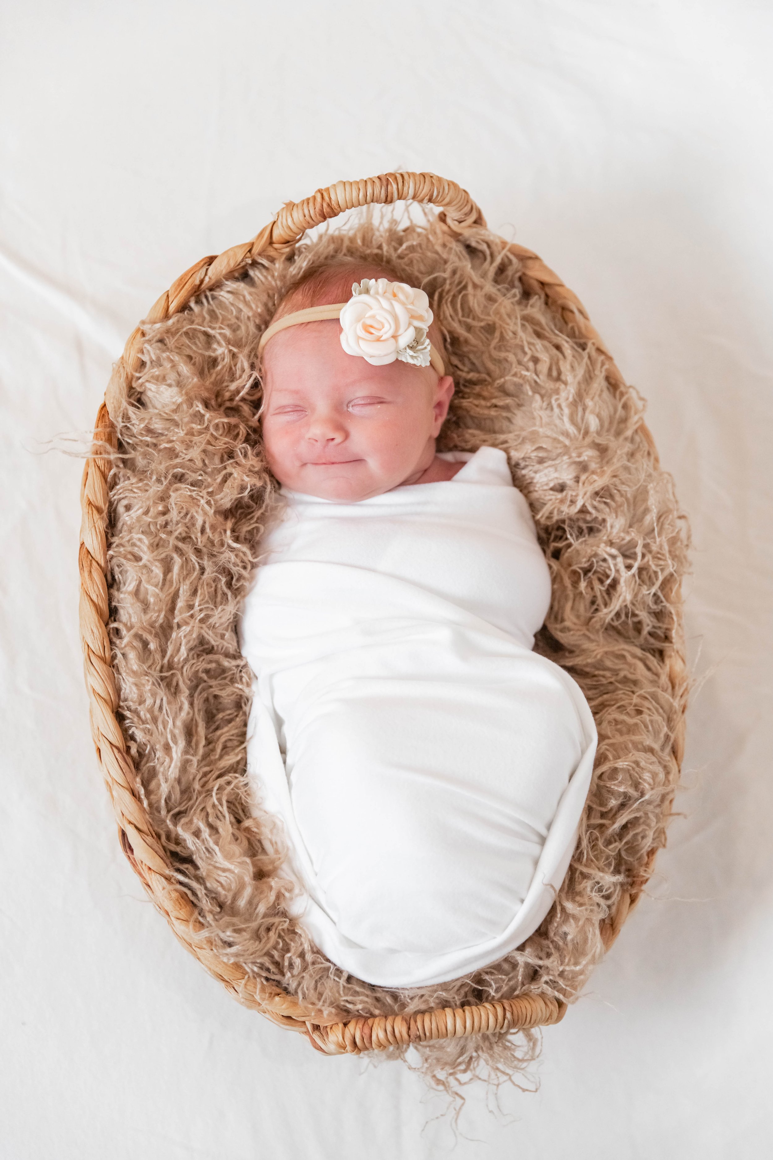 A smiling newborn baby wrapped in a white blanket lying in a wicker basket with a fur lining, wearing a beige headband with a white floral decoration.