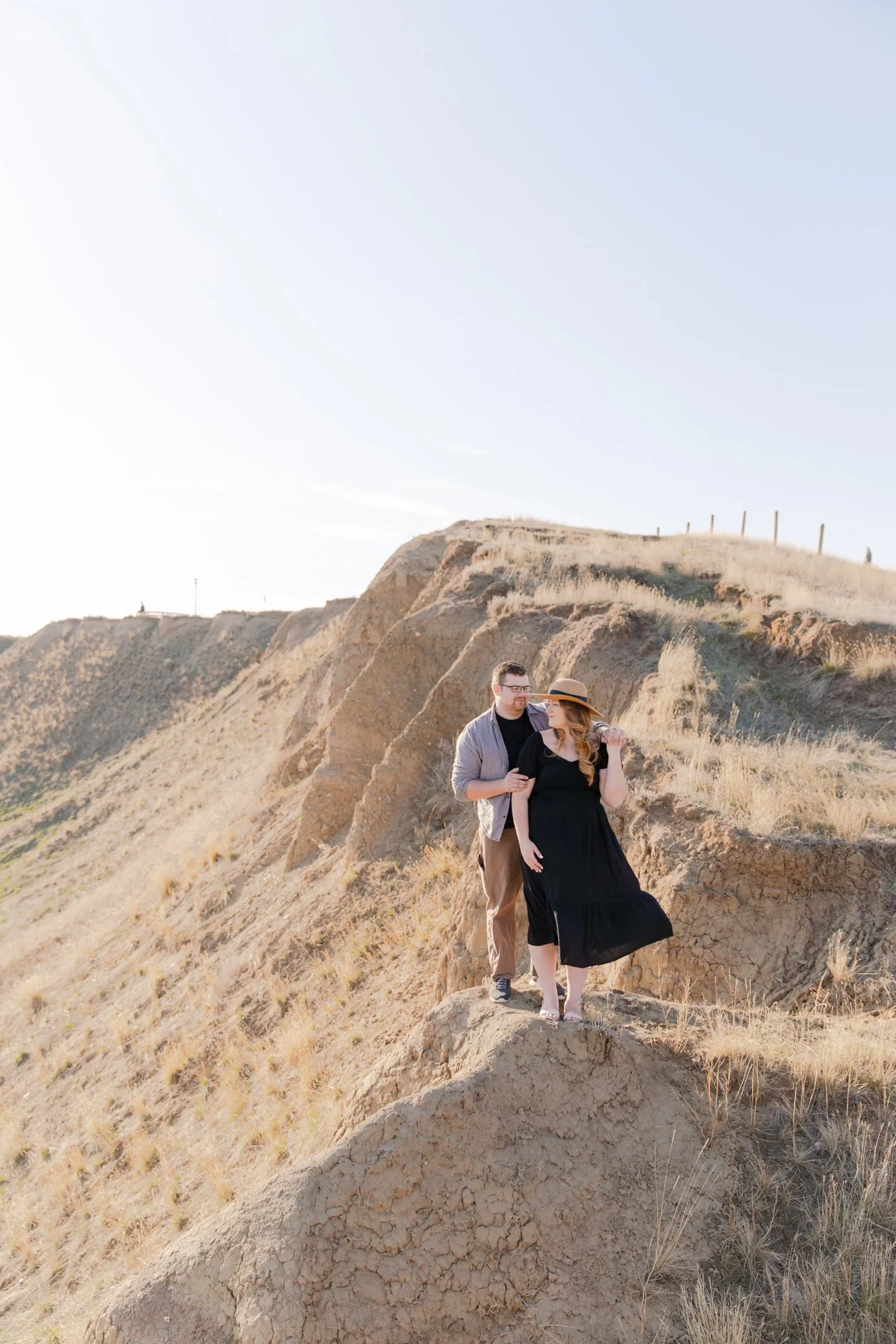 A couple standing on a rocky hill in a desert landscape, with the woman wearing a black dress and straw hat, and the man in glasses and casual clothing, during late afternoon sunlight.