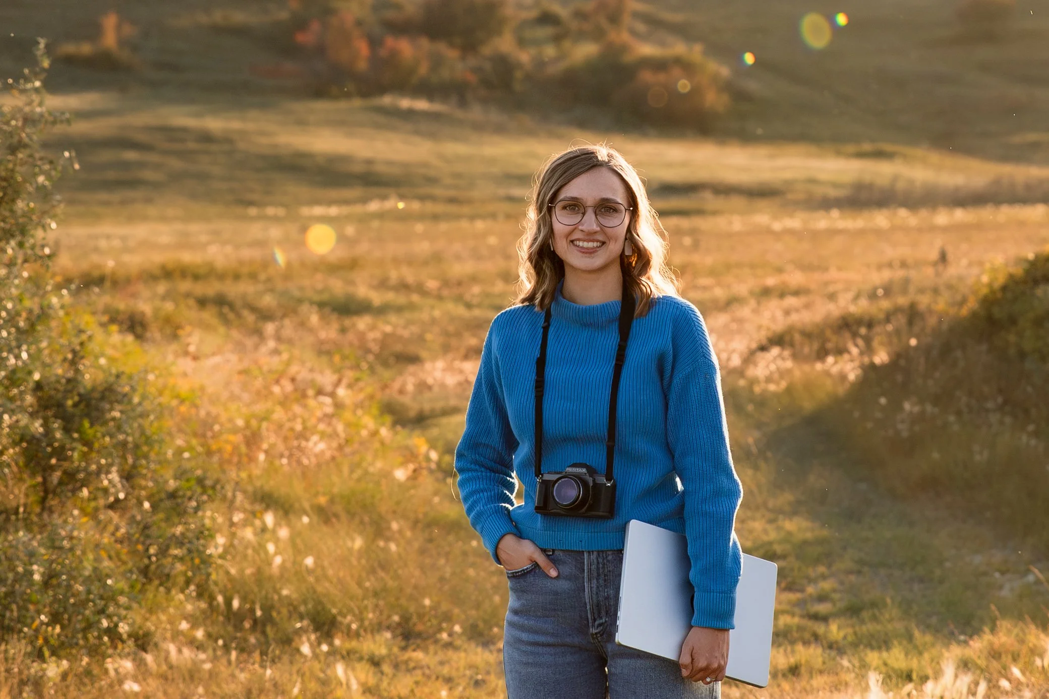 A smiling young woman with glasses and wavy hair standing outdoors in a field during sunset. She is wearing a blue sweater, jeans, and has a camera hanging around her neck. She is holding a white laptop in her left hand.