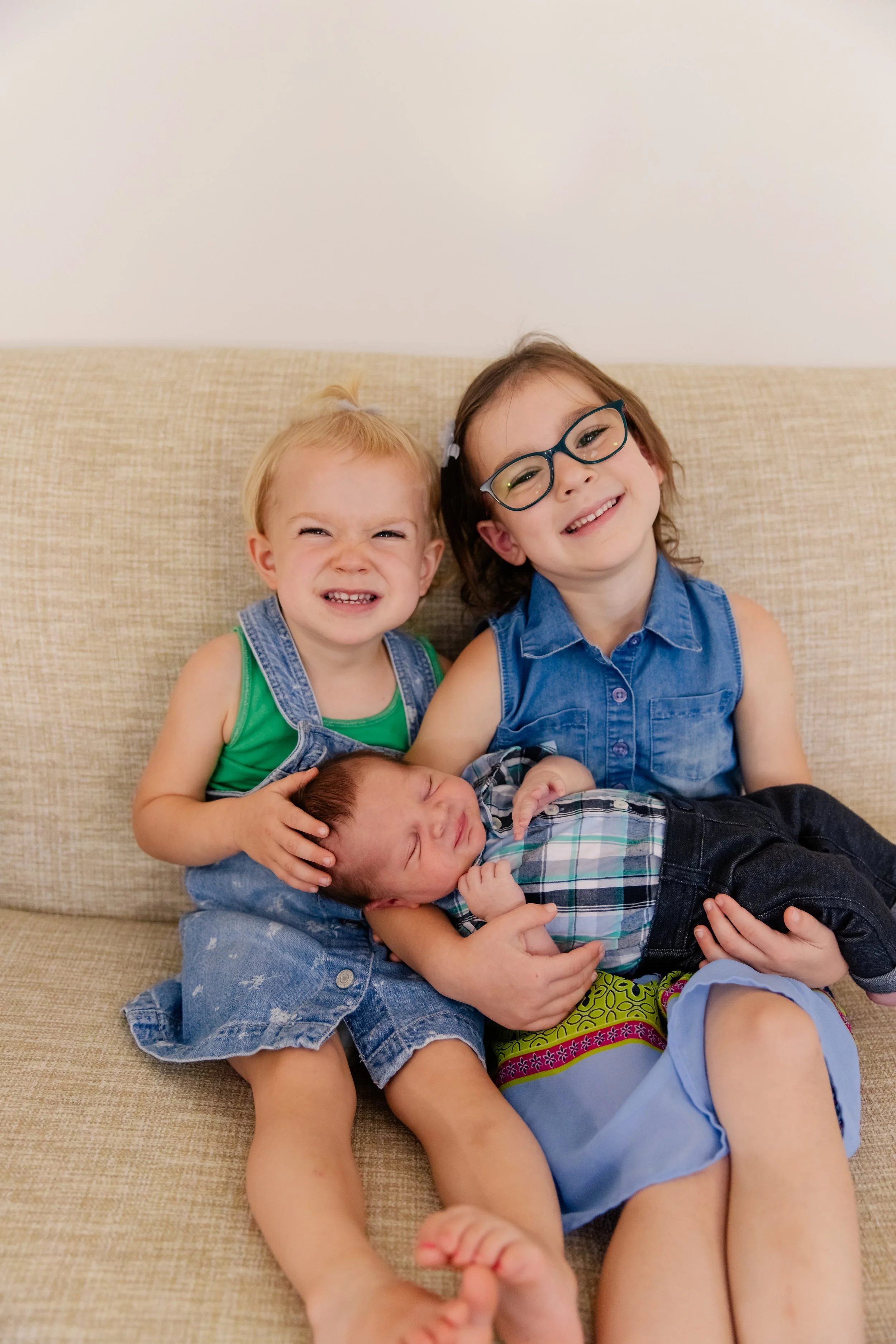Three children sitting on a beige couch, with a baby in the center. The baby is being held and appears to be crying. The girl on the right wears glasses and a blue sleeveless shirt, smiling. The girl on the left, in a green tank top and denim overalls, is making a face, also smiling.