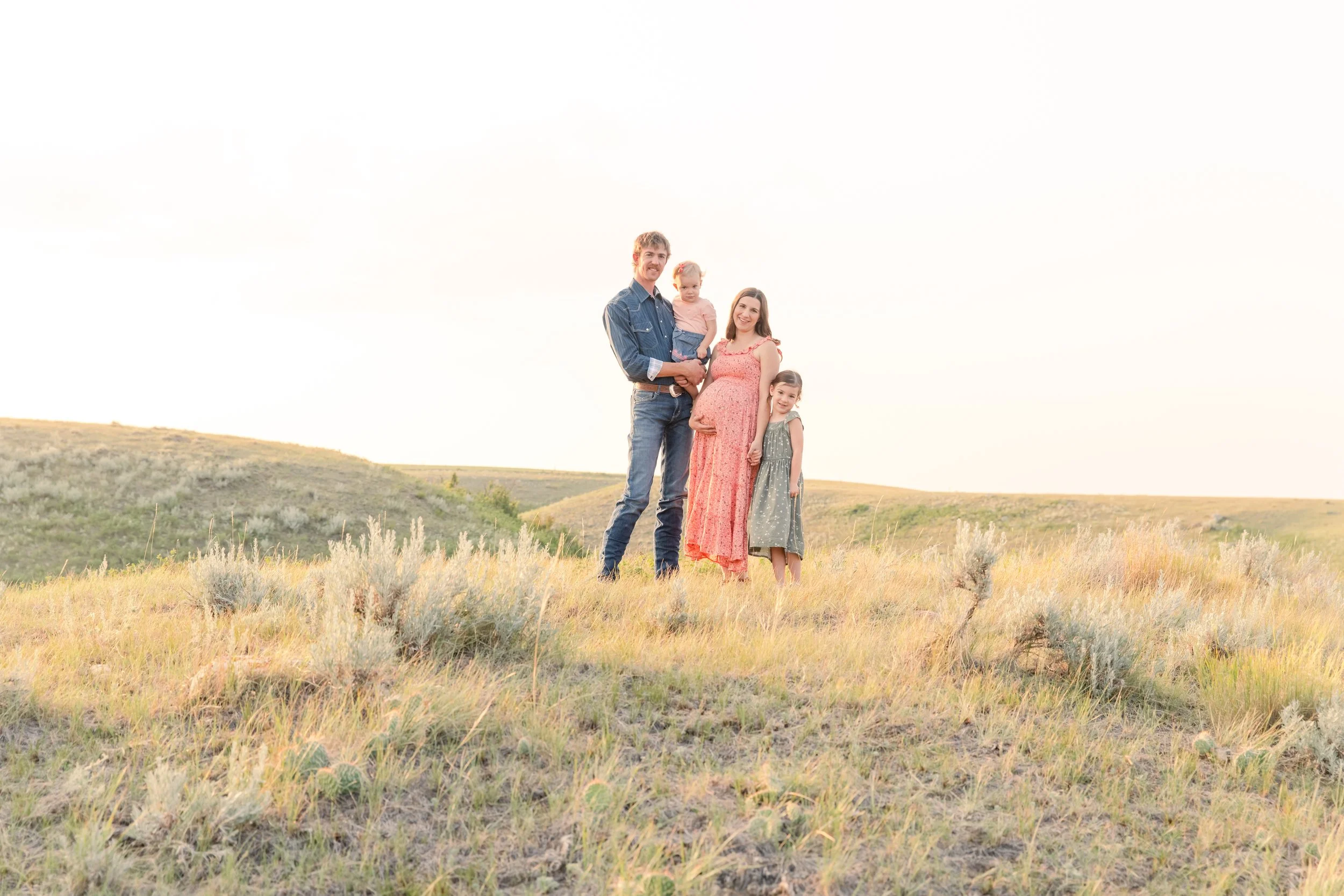 A family of four standing together in a grassy field with rolling hills in the background during sunset.