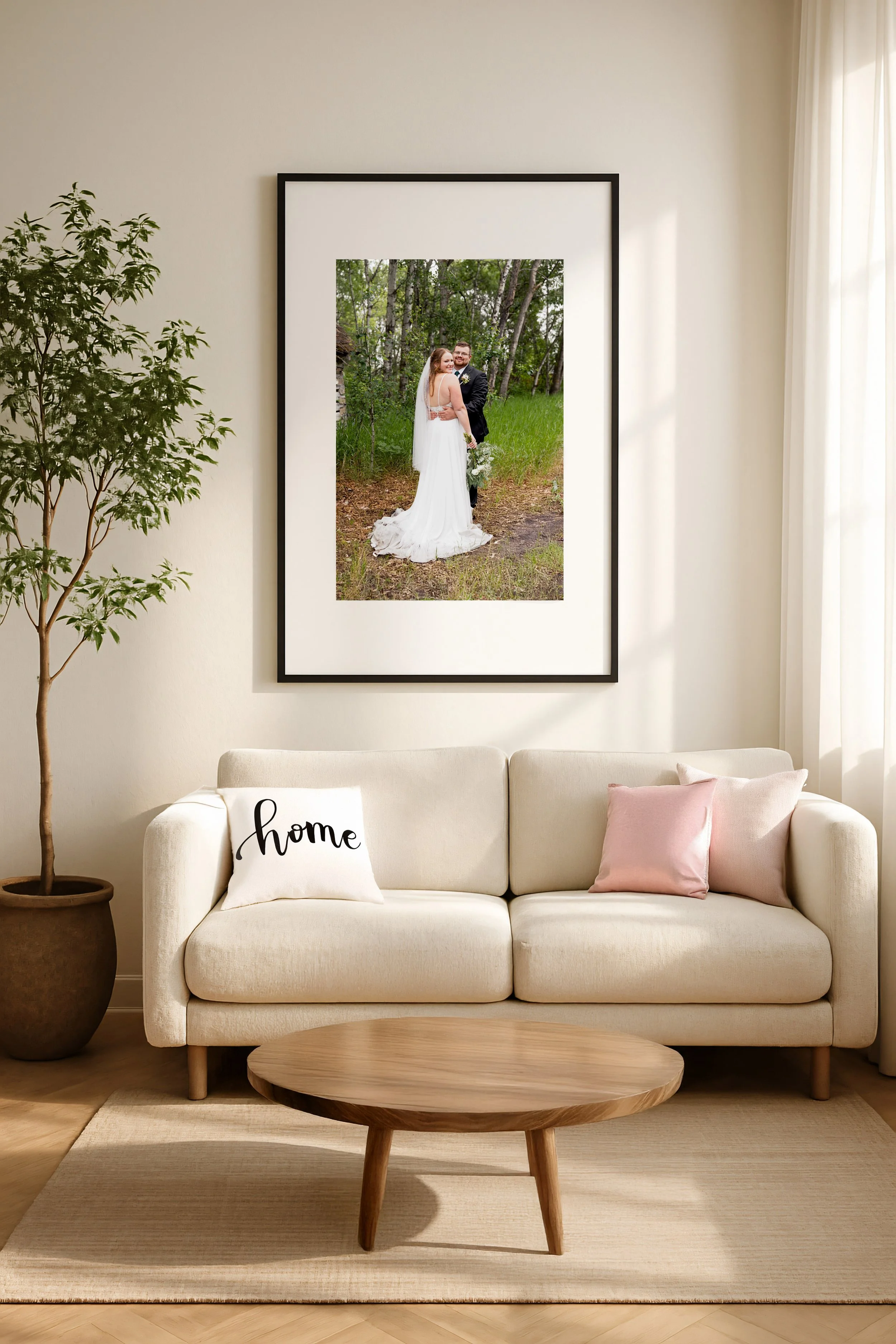 Living room with white couch, pink and white pillows, a round wooden coffee table, potted plant, and a large framed photograph of a bride and groom standing outdoors in a forest.