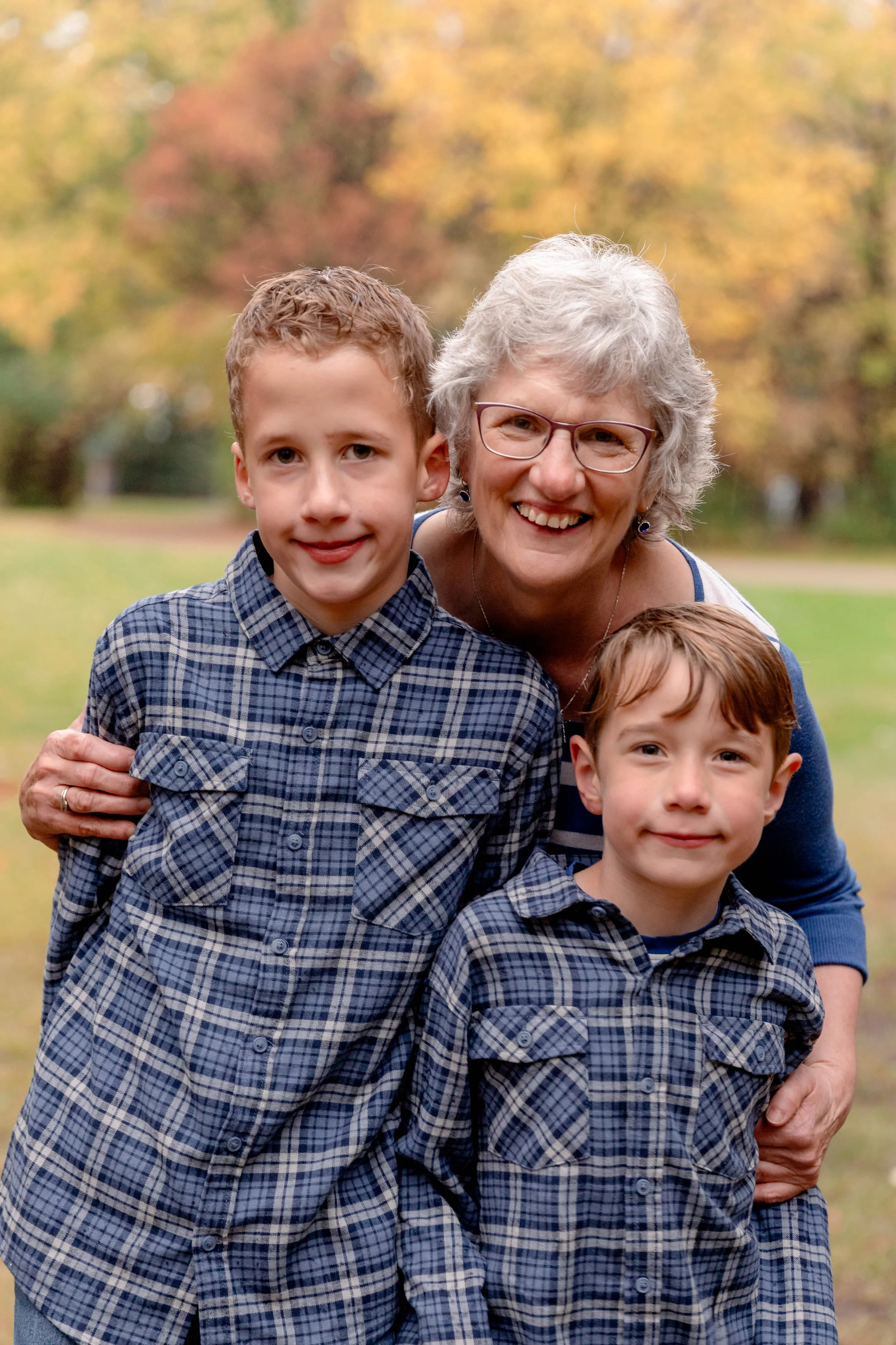 A smiling older woman with glasses and gray hair poses outdoors with two young boys dressed in plaid shirts. They stand on a grassy area with colorful autumn trees in the background.