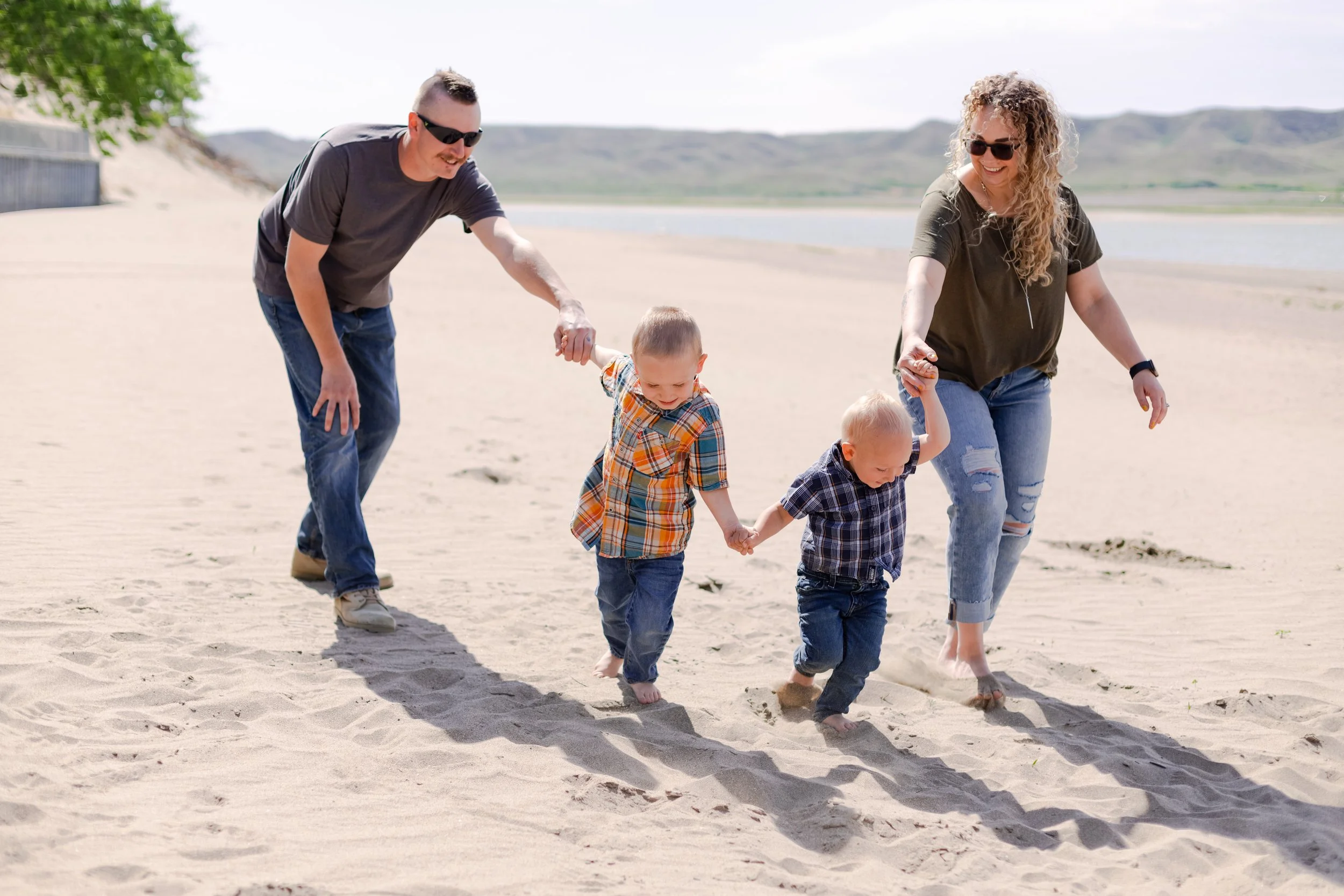 A family of four walking barefoot on the beach, holding hands and enjoying the sunny day with a lake and hills in the background.