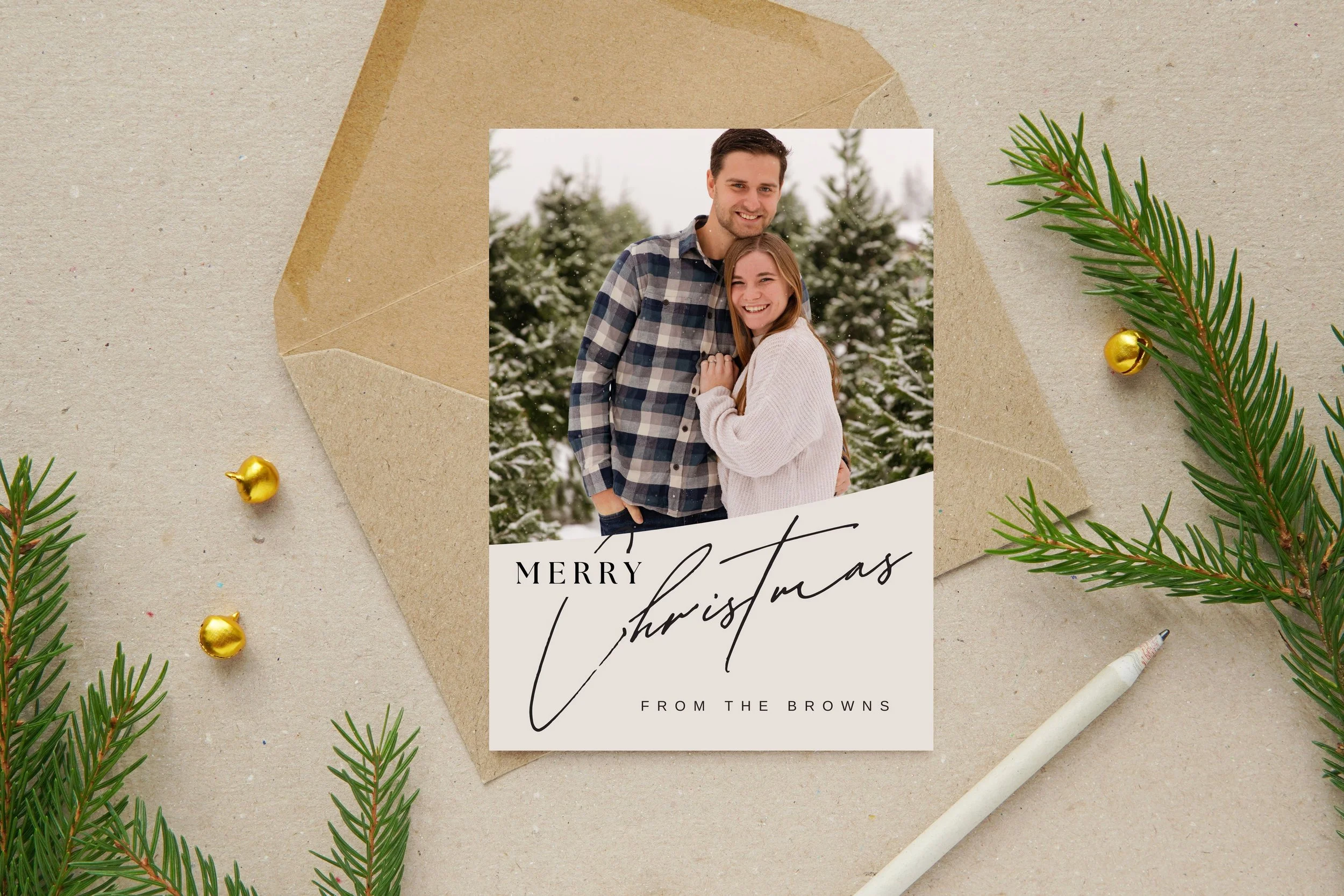 A Christmas greeting card featuring a smiling couple in winter clothing, standing outdoors with snow-covered trees in the background. The card reads, "Merry Christmas from the Browns." The card is placed on a beige surface decorated with evergreen branches, small gold ornaments, a white pen, and a brown envelope.