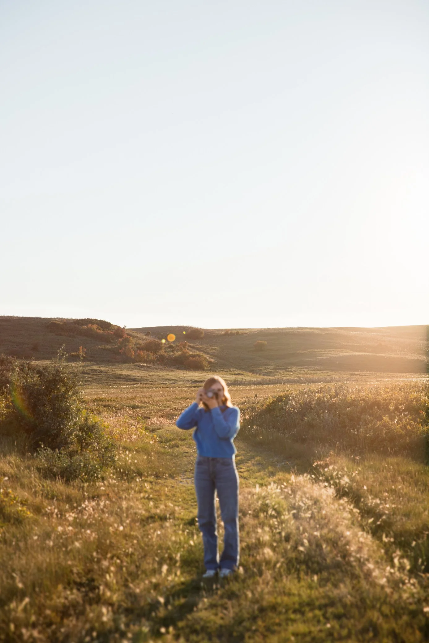 A person standing on a grassy trail in a wide open field, taking a photo with a camera in the late afternoon or early evening sunlight.