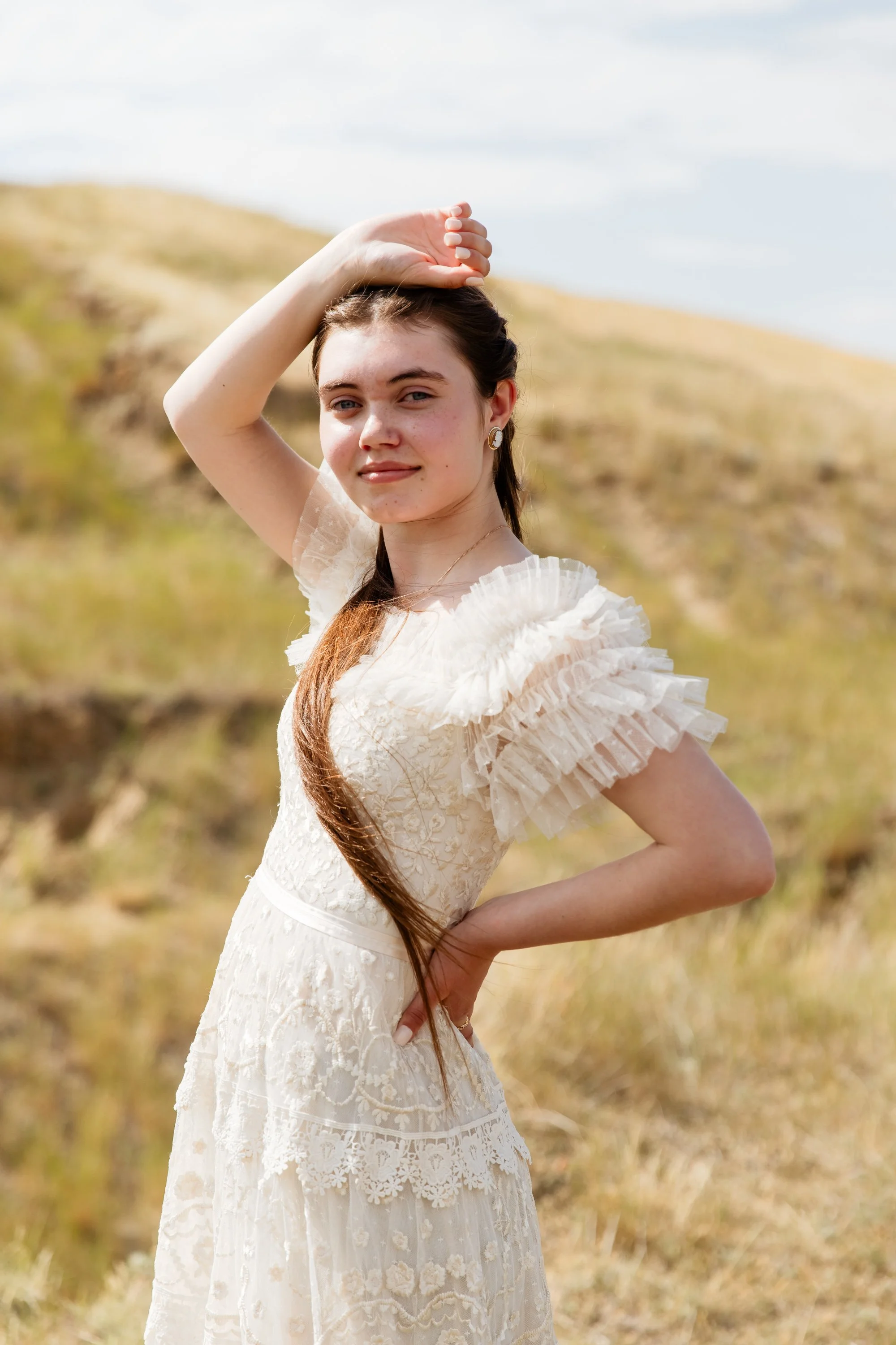 A young woman in a white lace dress with ruffled sleeves standing outdoors in a grassy landscape, posing with her right hand on her hip and her left hand touching her head, with long brown hair.